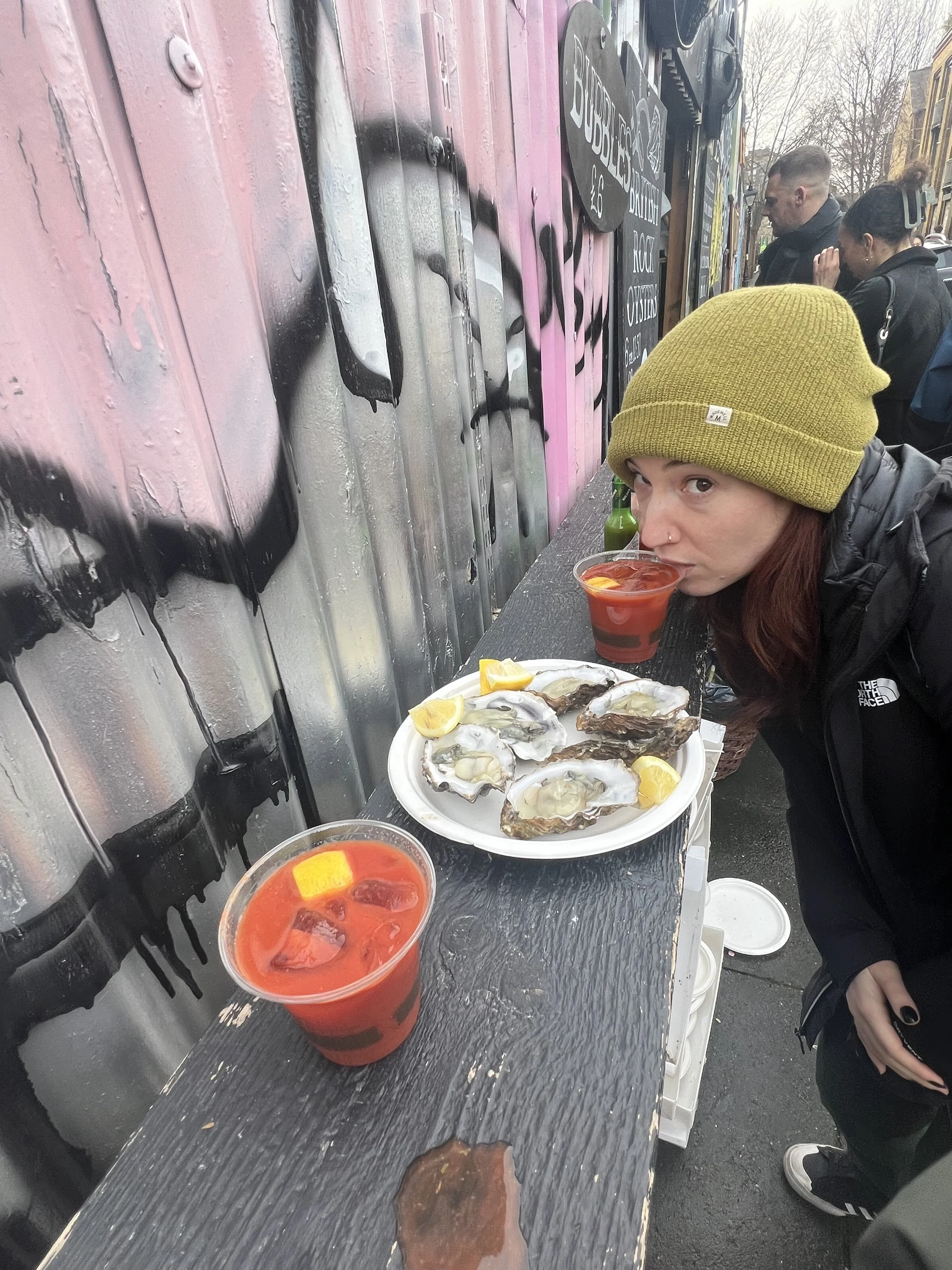 A woman wearing a green beanie and black jacket leaning over a black table with fresh oysters on a white plate, lemon wedges, a red cocktail with ice and lemon, and a green bottle outside in an urban setting.