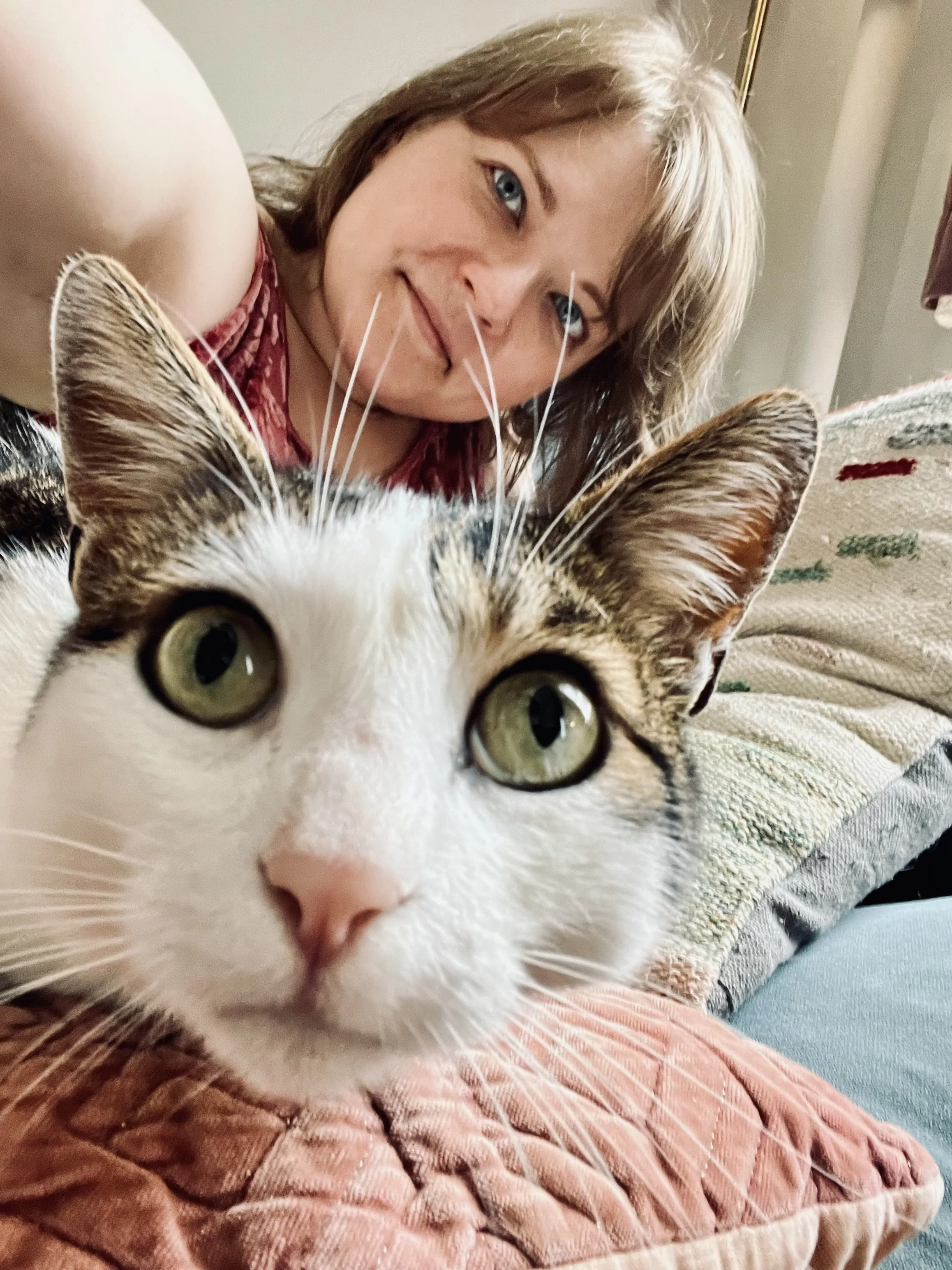 A woman with blonde hair and blue eyes taking a selfie with a close-up view of her tabby and white cat, both lying on a textured pink and orange quilted blanket on a couch.