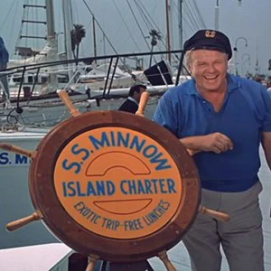 Smiling man in a blue shirt and navy cap standing behind a boat's wheel with a sign reading 'S.S. Minnow Island Charter' in a marina.
