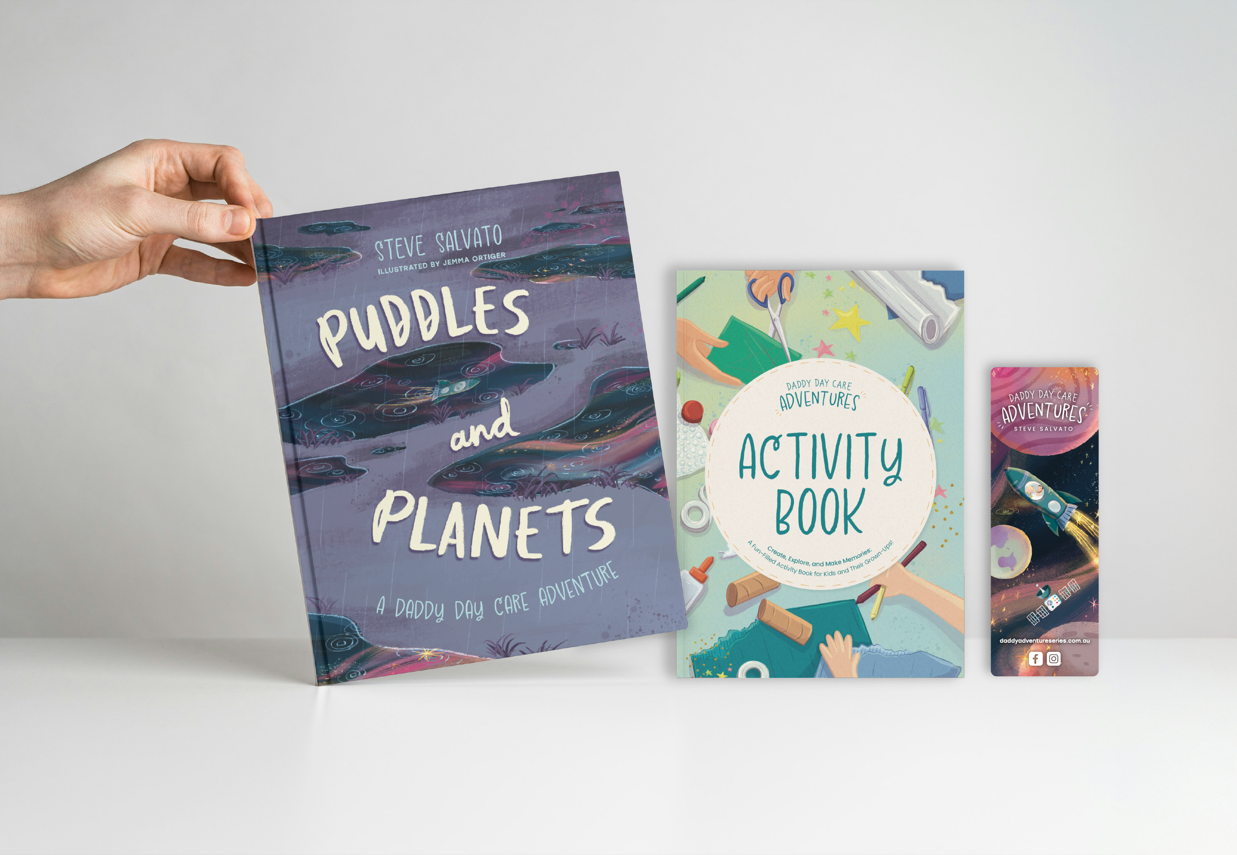 A person holding a children's book titled "Puddles and Planets" by Steve Salvato, with illustration by Jemma Ortiger. Other colorful activity books about adventures and activities for kids are displayed on a white table.