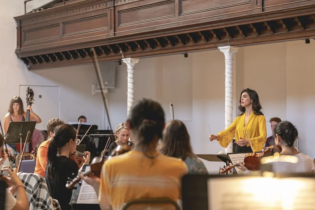 Orchestra rehearsing with a woman conductor in a yellow blazer leading the musicians in a classroom or hall with a wooden balcony.