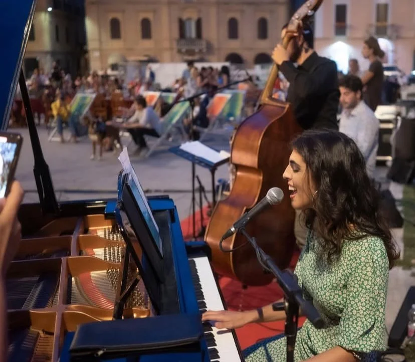 A woman singing and playing piano, with a man playing double bass behind her, during an outdoor jazz performance at night with an audience in the background.