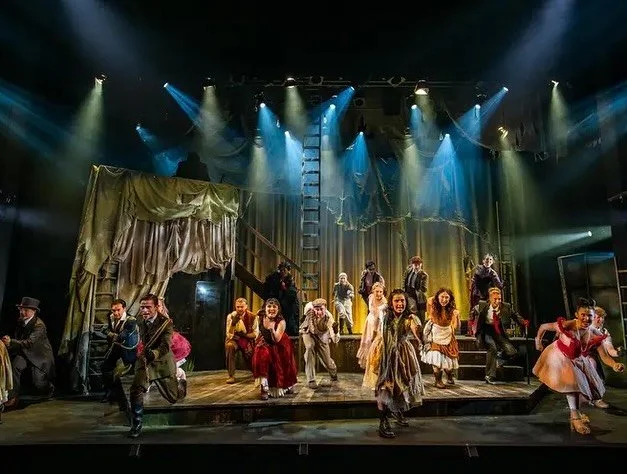 Theater stage with performers in period costumes under colorful stage lighting, with a multi-level set and staircase.