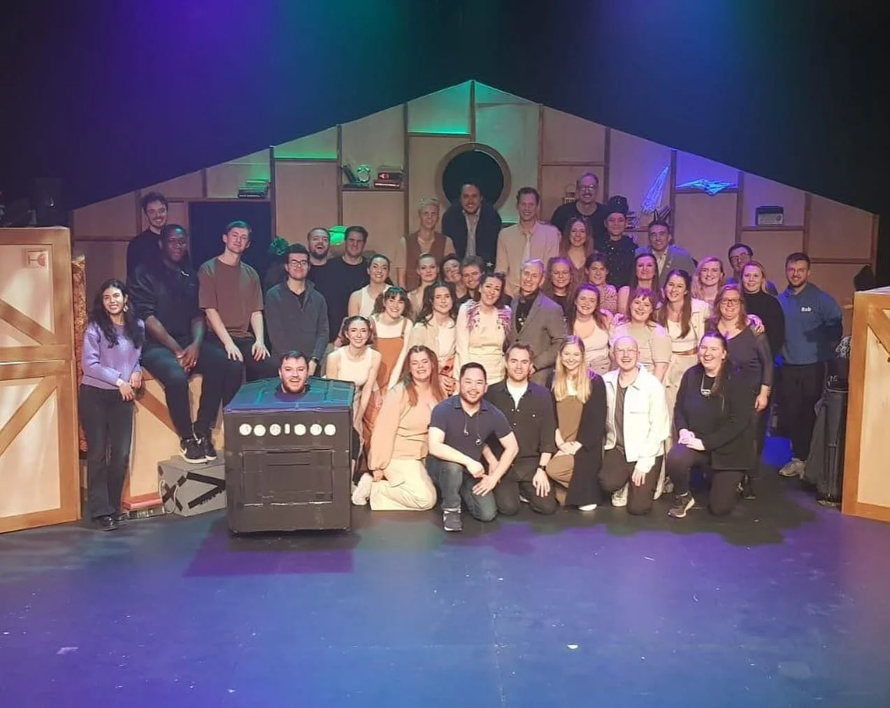 Group photo of theater cast and crew on stage with set design in the background.