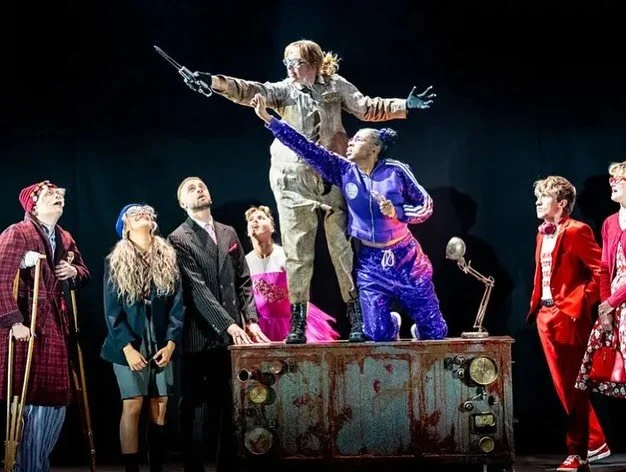 Group of performers on stage, with two women standing on a rusty, vintage radio, one reaching out with a sword, surrounded by others in colorful costumes, under bright stage lighting.