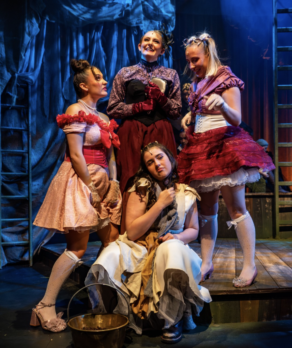 Five women in colorful vintage costumes on stage during a theatrical performance, with blue stage curtains in the background.