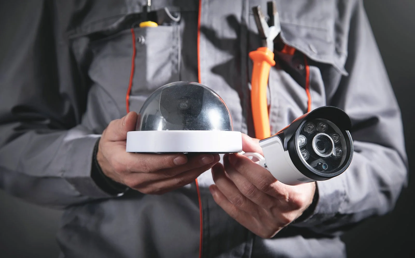 Person in gray work jacket holding a dome-shaped security camera with a white base and a black, circular lens module.