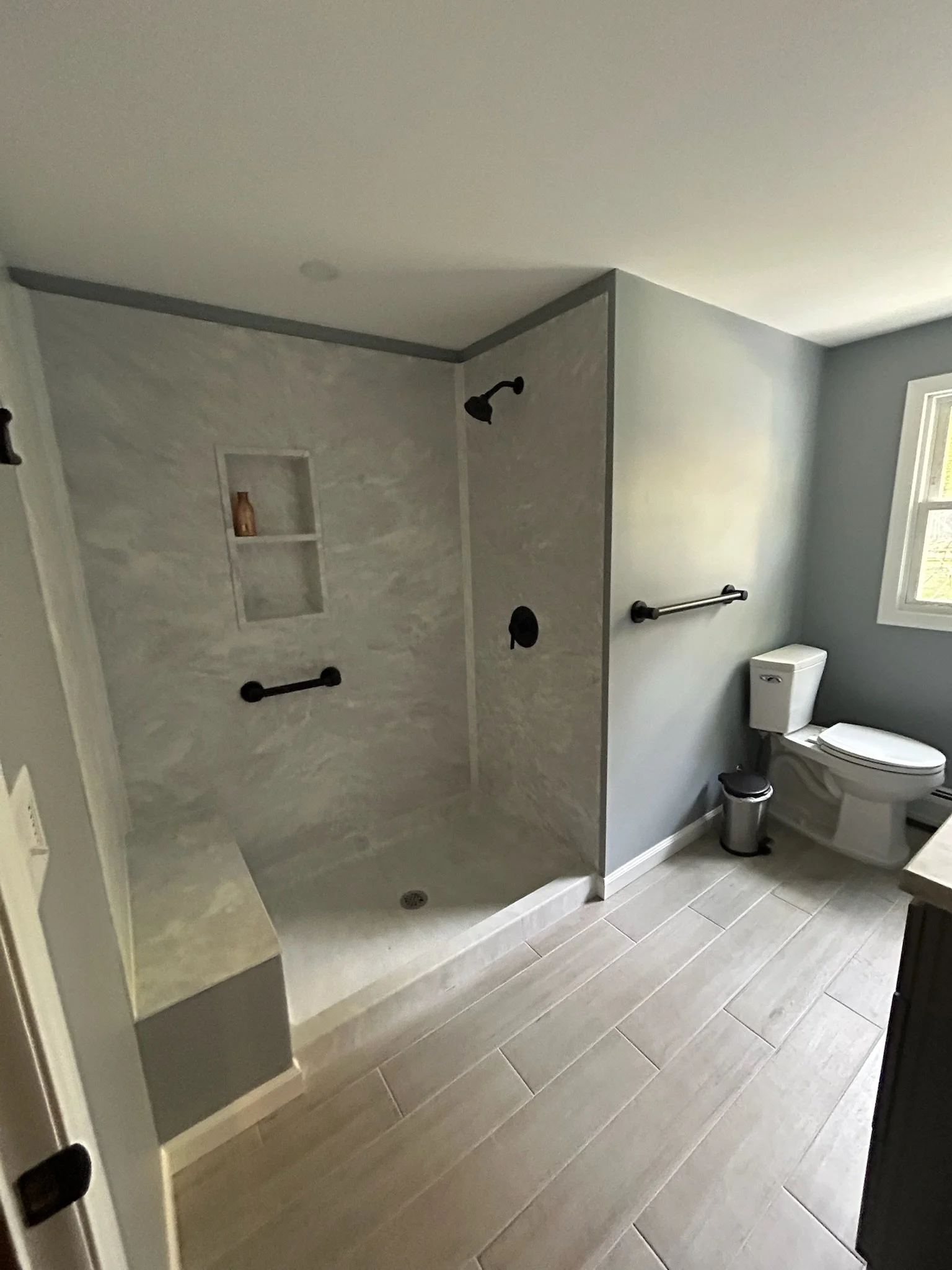 Bathroom with a walk-in shower with gray marble walls and black fixtures, a built-in niche, a toilet, a small waste bin, a window, and light-colored tiled flooring.