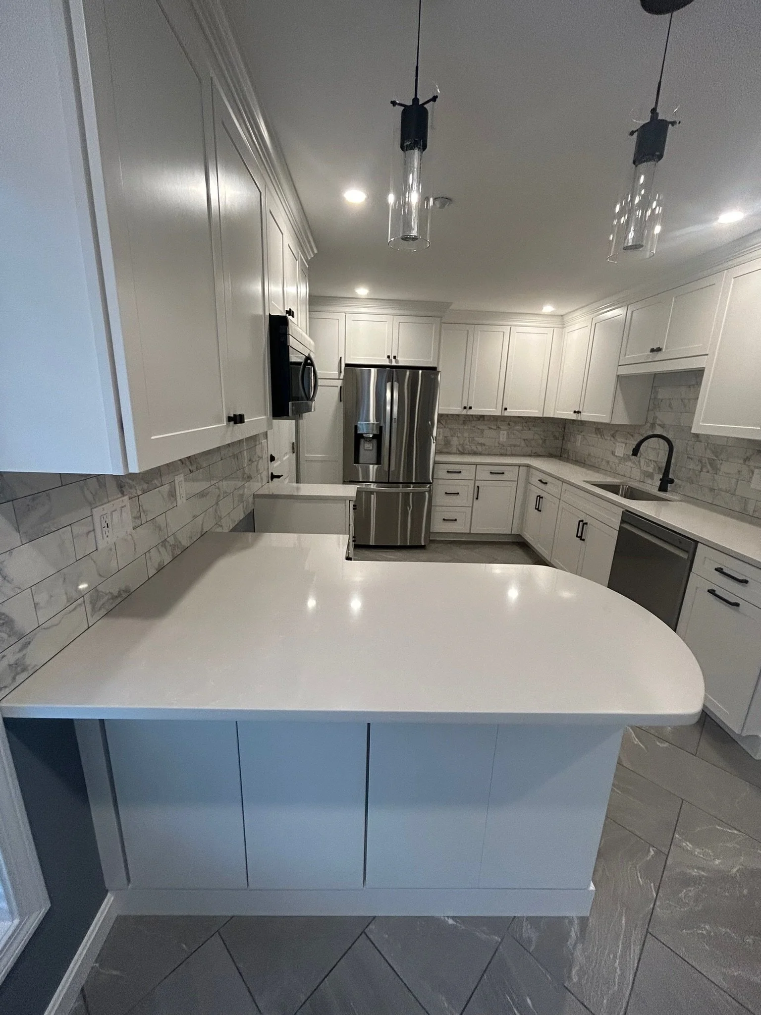 Modern kitchen with white cabinets, marble backsplash, stainless steel appliances, and a white countertop island, illuminated by ceiling lights and pendant lights.