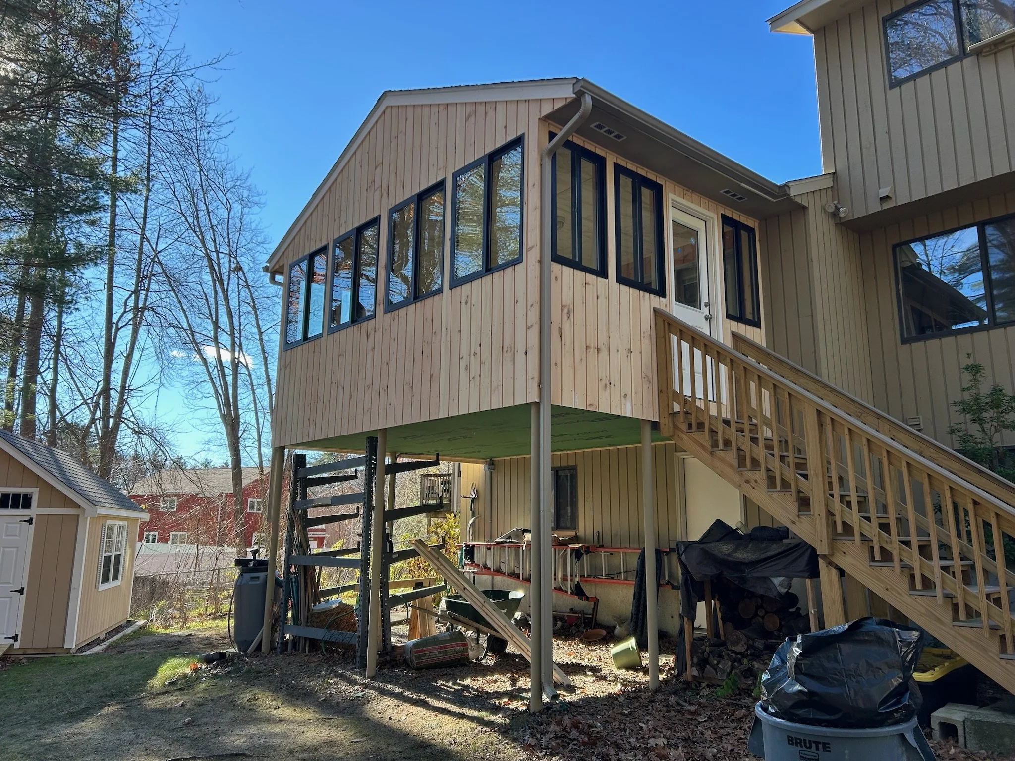 A two-story house with a wooden extension or sunroom added on the upper level, accessed by an outdoor staircase, located in a backyard with trees and other structures nearby.