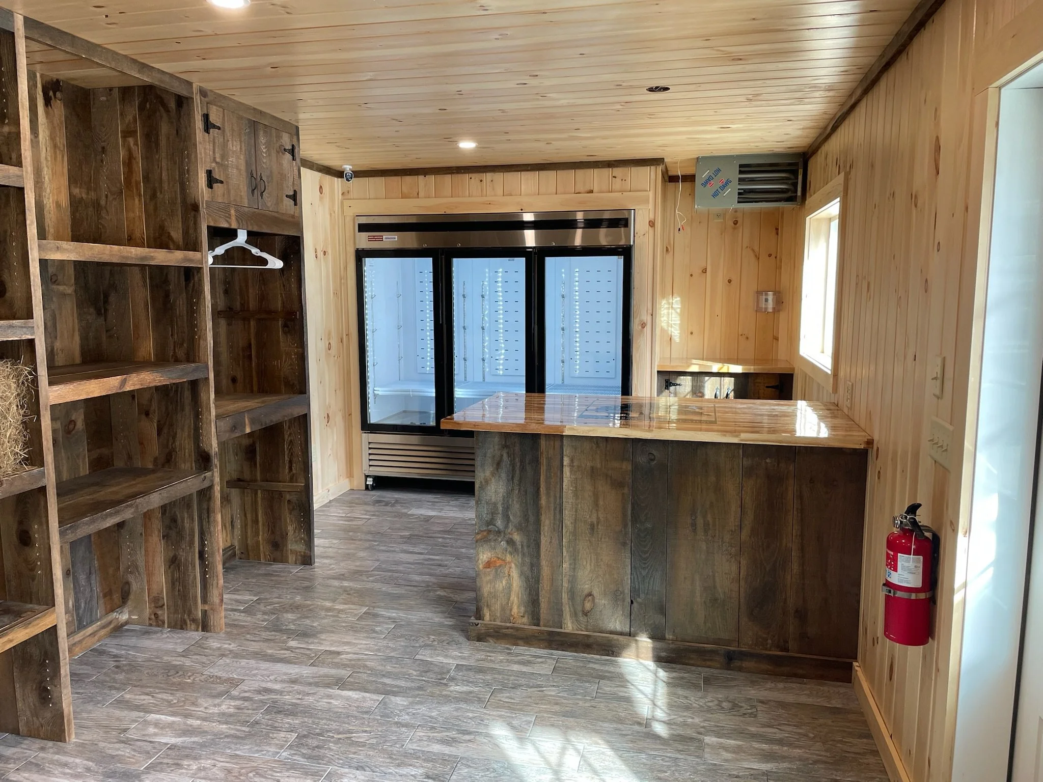 Interior of a rustic wooden room with shelves, a counter, and a refrigerator, featuring natural wood paneling and flooring.