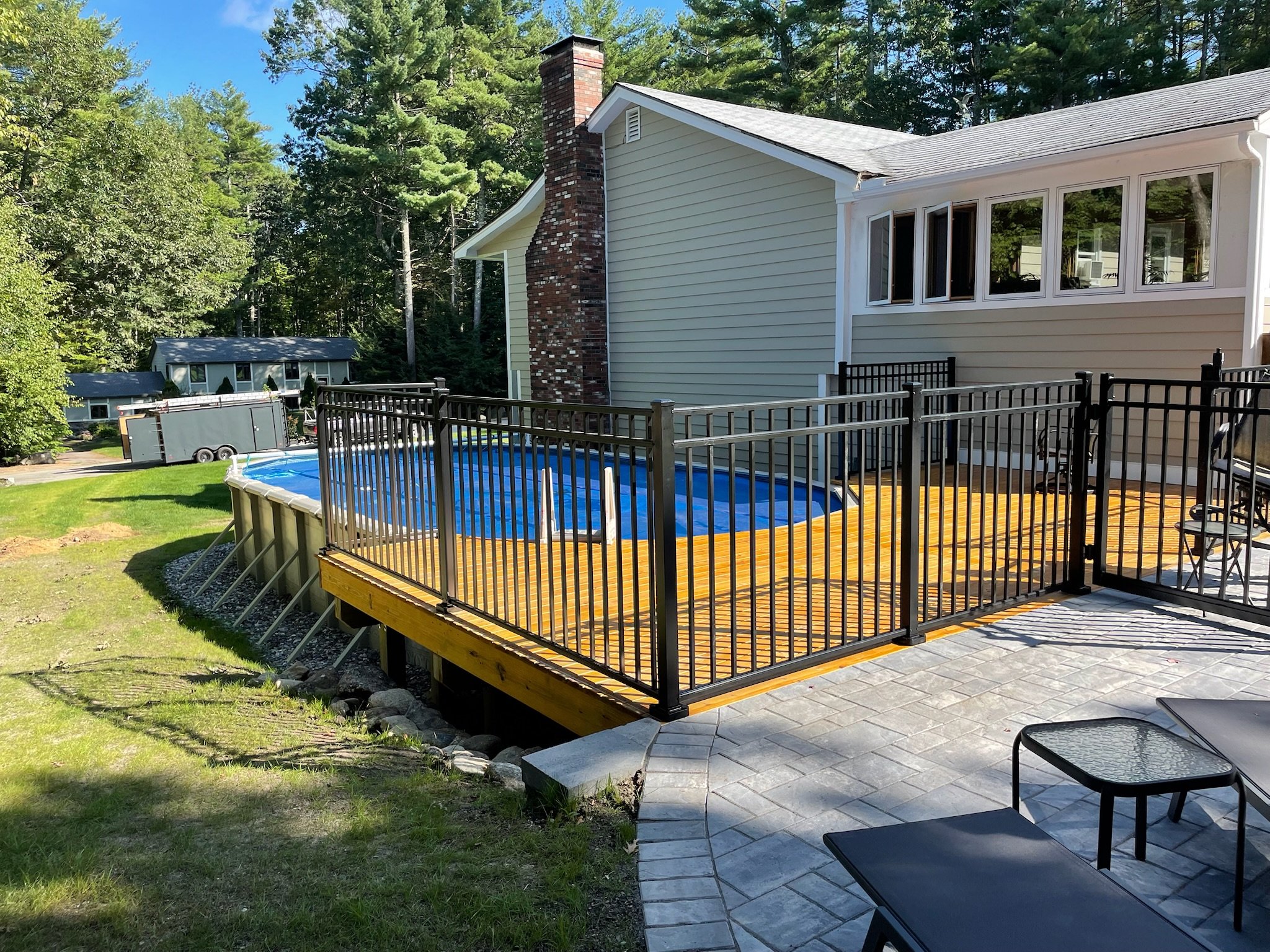 Residential backyard with a wooden deck and above-ground pool, fenced with black metal railing, adjacent to a house with beige siding and a brick chimney, surrounded by green trees.