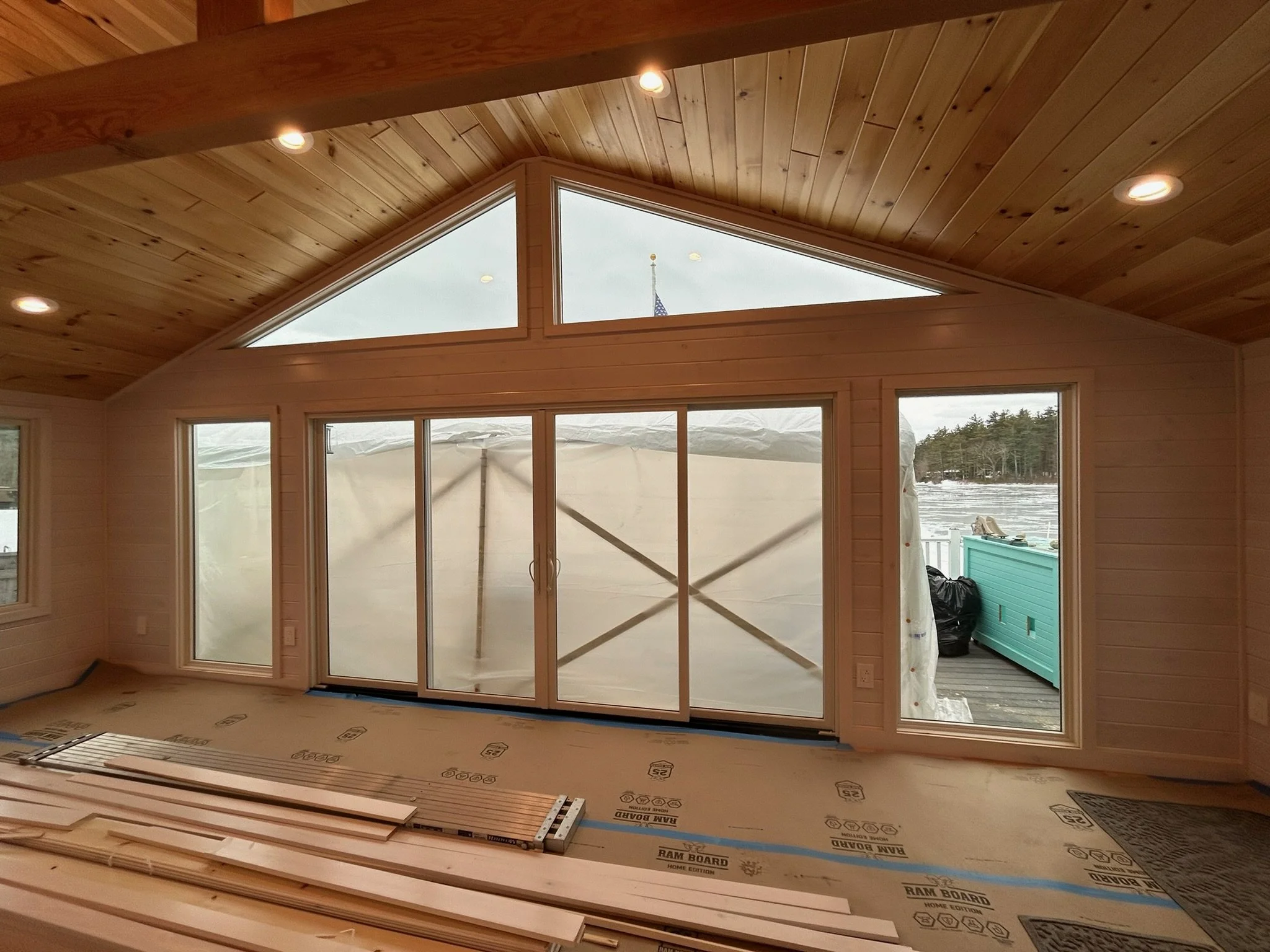 Interior of a house under construction with a wooden ceiling, large sliding glass doors, and windows, overlooking a snowy outdoor landscape.