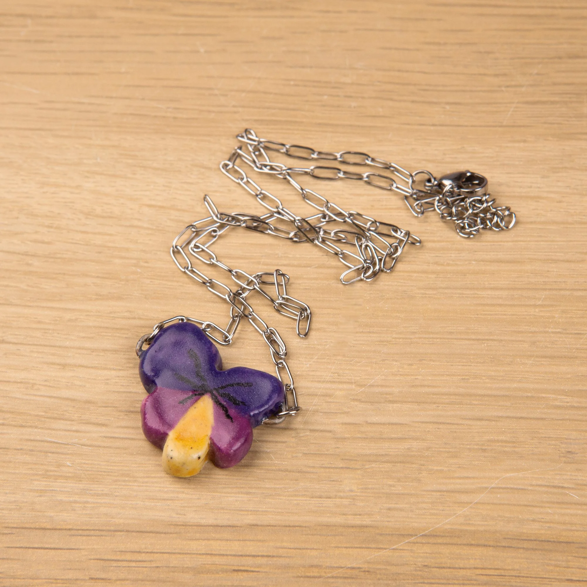 Handmade ceramic pansy necklace featuring a playful hand-painted pansy pendant on a silver-coloured chain.