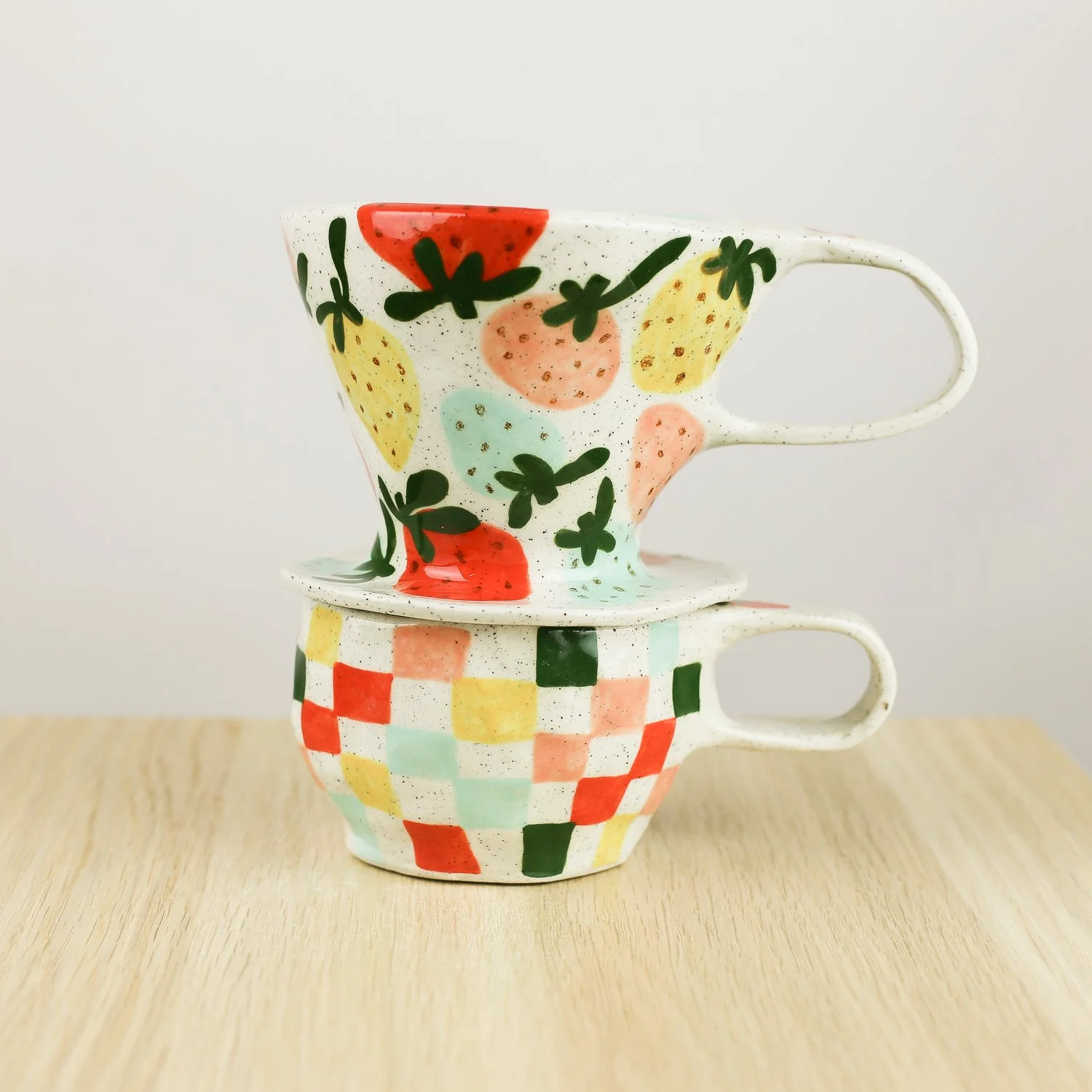 Handmade ceramic strawberry coffee dripper placed on a strawberry check cup, styled together on a soft, light background