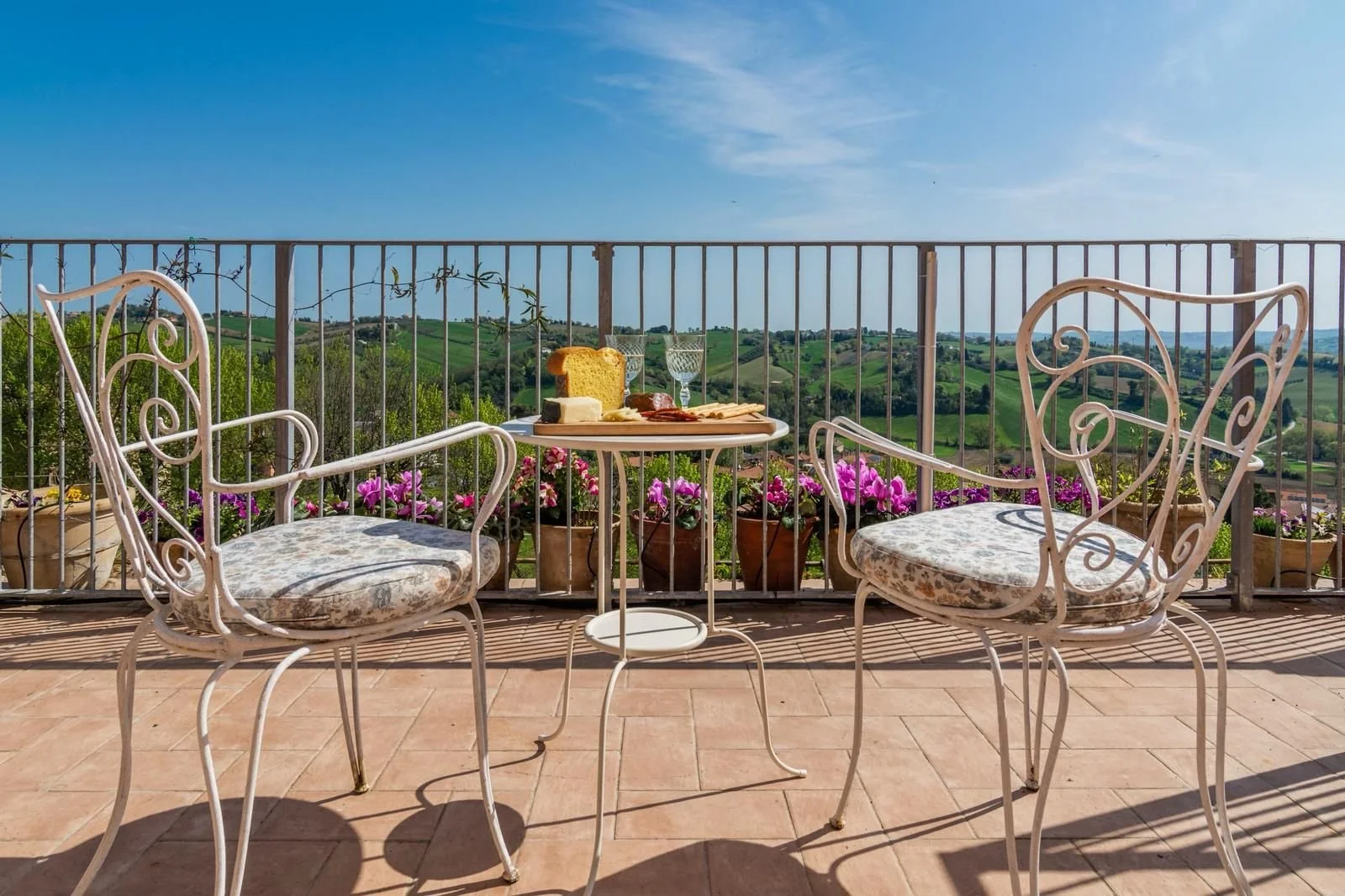 A scenic outdoor patio overlooking rolling green hills on a bright sunny day, with a small round table set for breakfast and two ornate white chairs with floral cushions, surrounded by pink flowering plants.