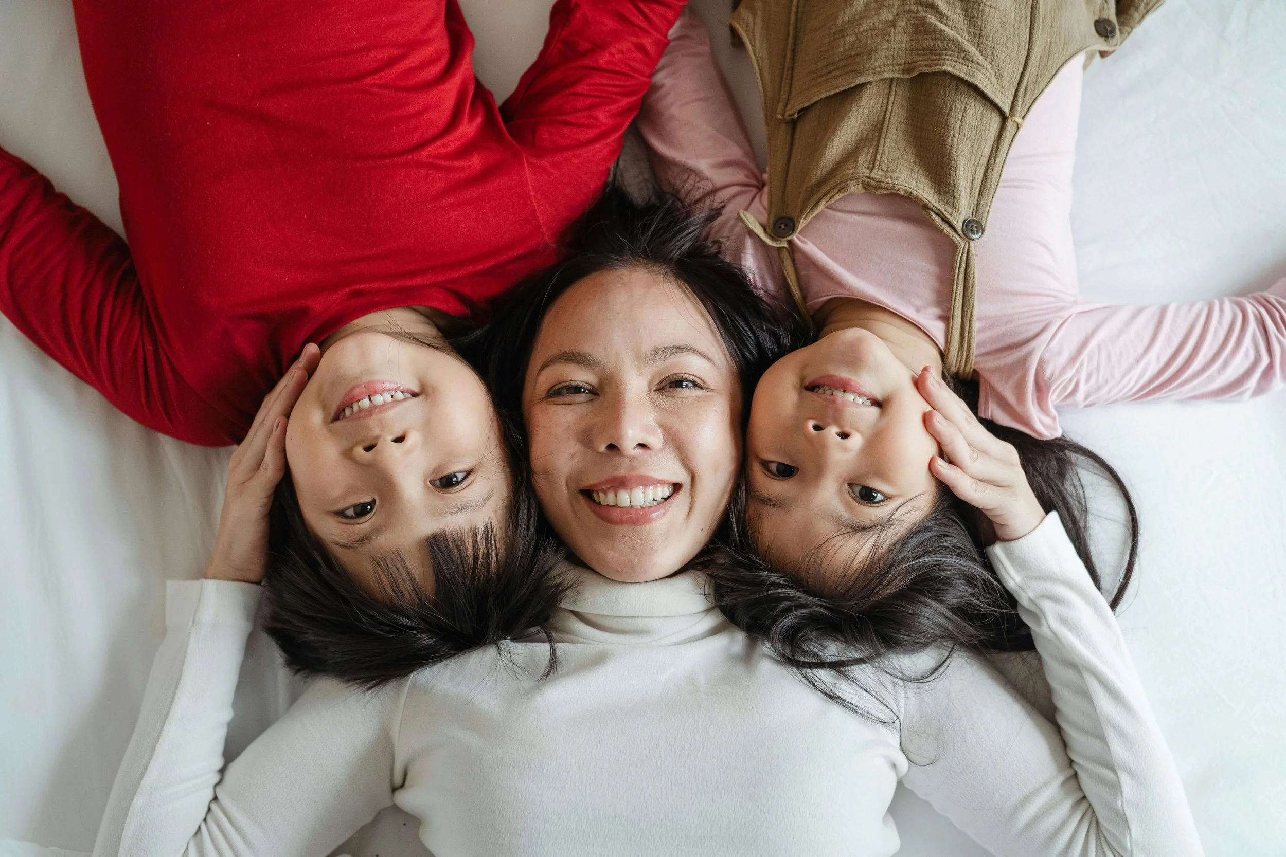 A smiling mother lies between her two young daughters. Is your family navigating the tensions between cultural heritage and the pressures of life in America? AAPI family therapy in Arcadia, CA, can help you communicate across generations.