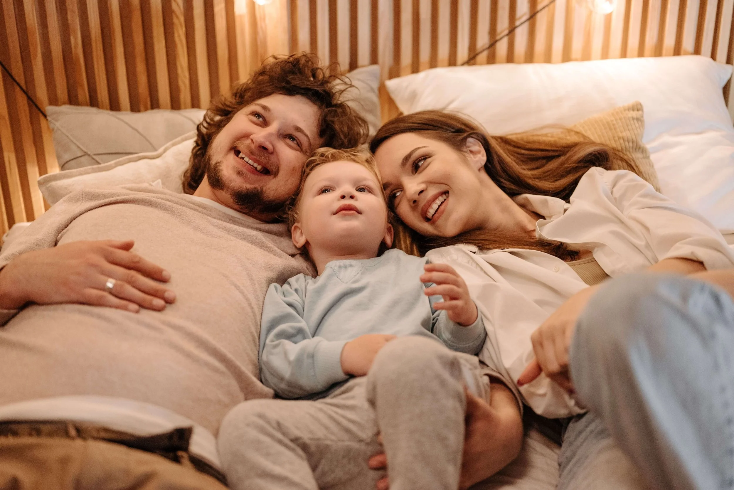 A smiling father, mother, and young son lie in bed together.