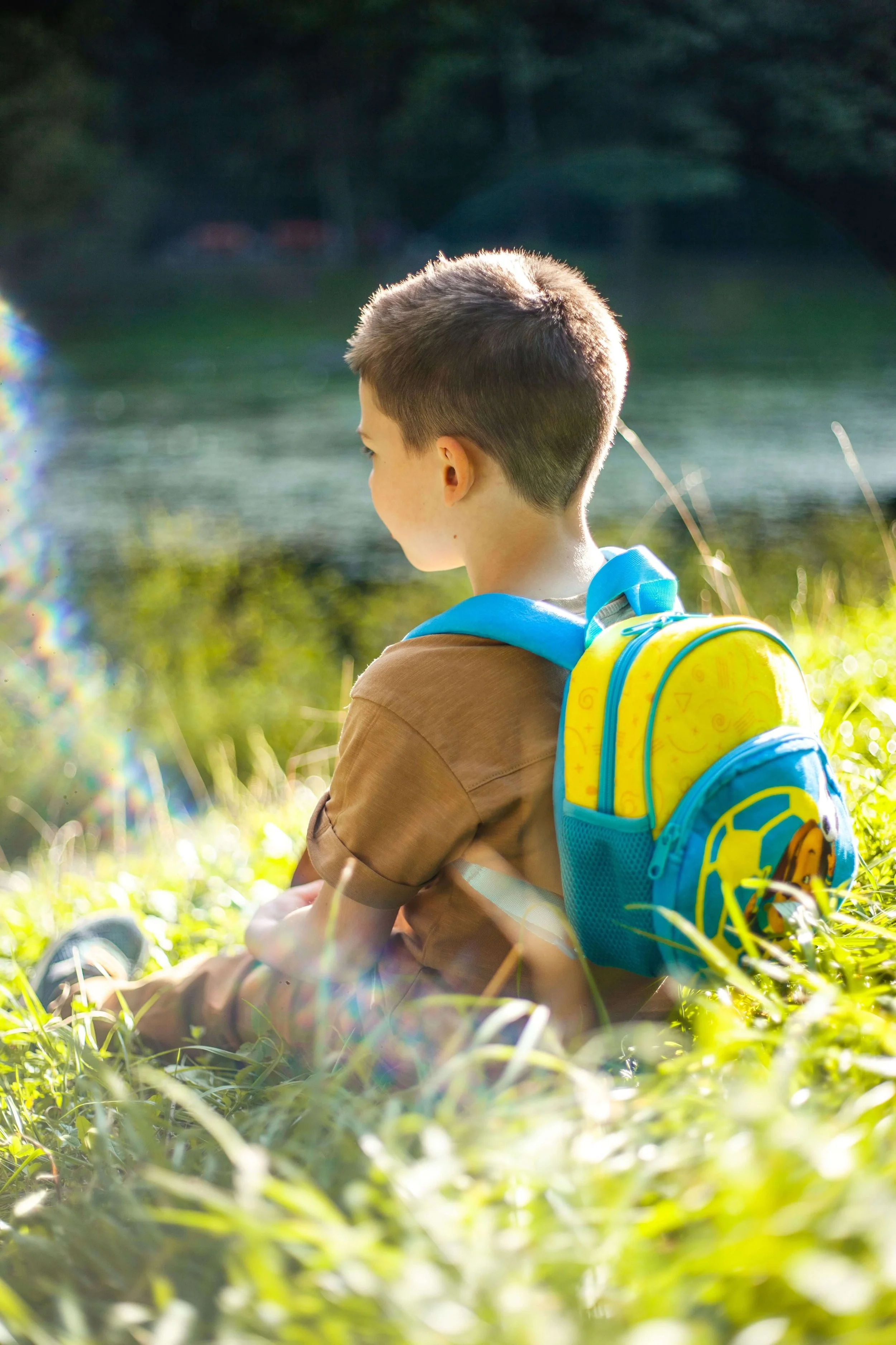 A child wearing a blue and yellow backpack sits in the grass outside.