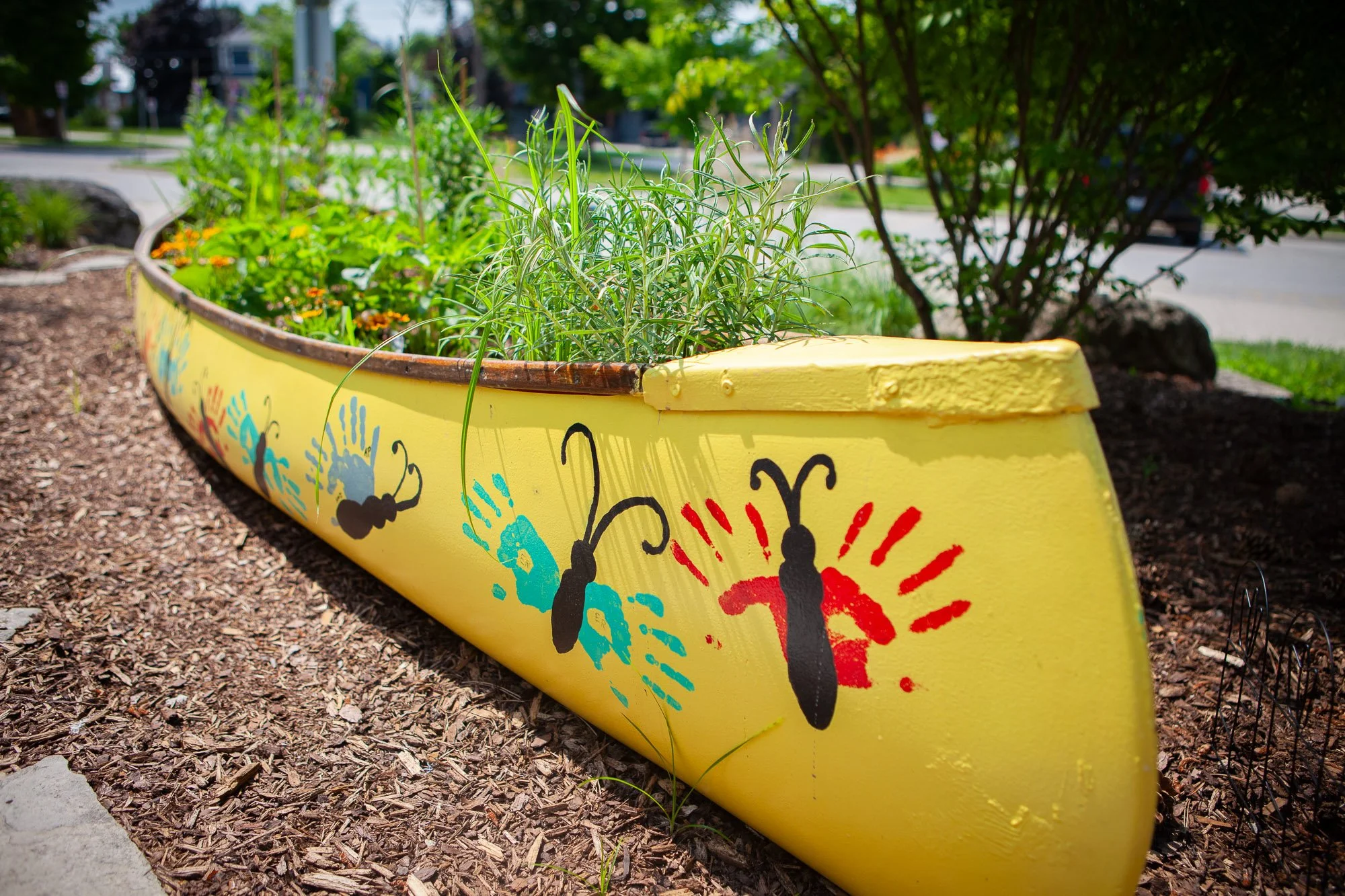 A yellow painted boat-shaped flower bed with colorful handprint and butterfly decorations, filled with green plants, on a garden bed with mulch, outside in a neighborhood with trees and houses in the background.