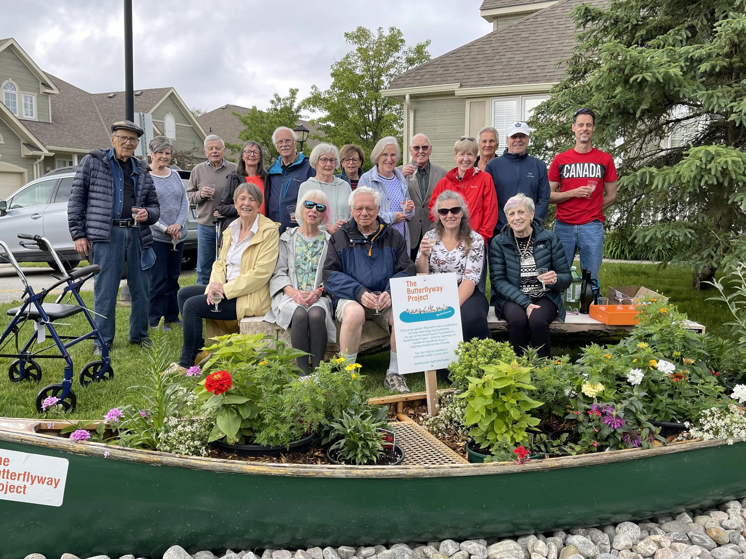 A group of people posing outdoors in a residential area next to a canoe filled with flowers and plants. A sign for The Butterflyway Project is visible. The group appears to be enjoying a gathering with multiple people holding drinks. The background includes houses, trees, and a parked car.