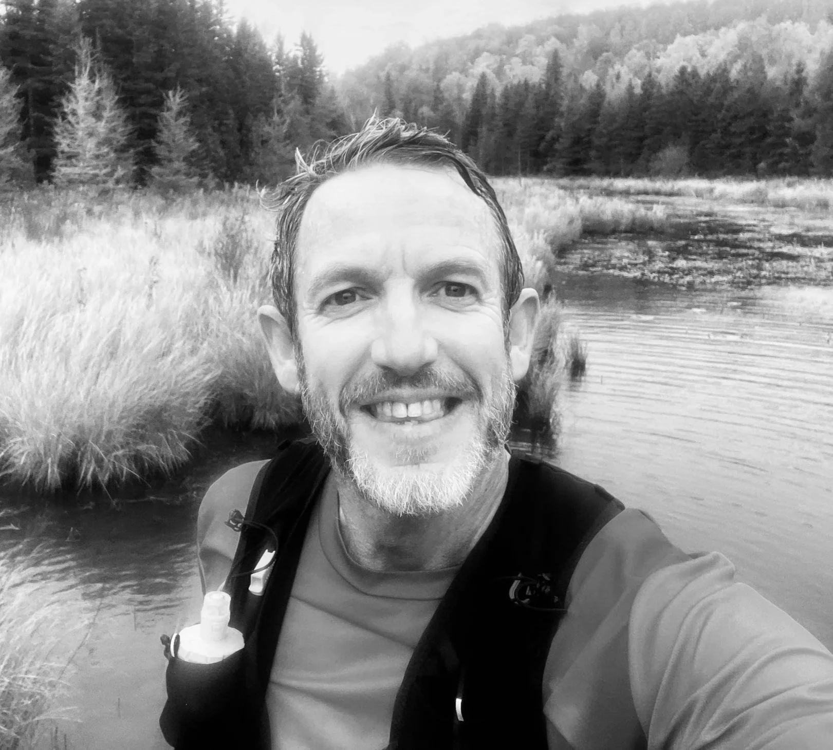 A man wearing outdoor gear, smiling, taking a selfie in a natural setting with a body of water, grasses, and forested hills in the background.