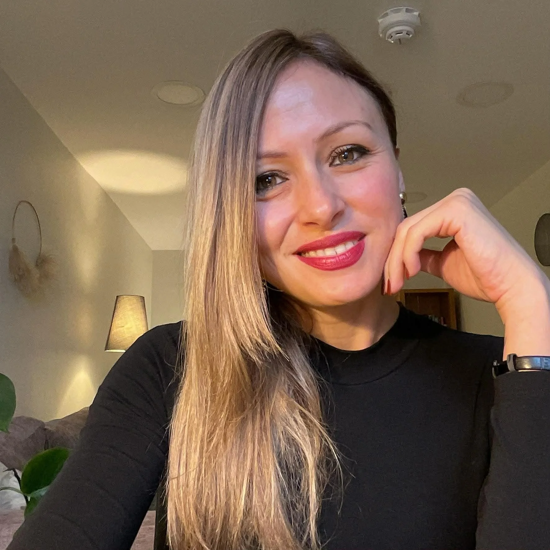 This image is of Neliona Moutsou a registered therapist with the BACP,  smiling with her left hand near her face, in a warmly lit room.