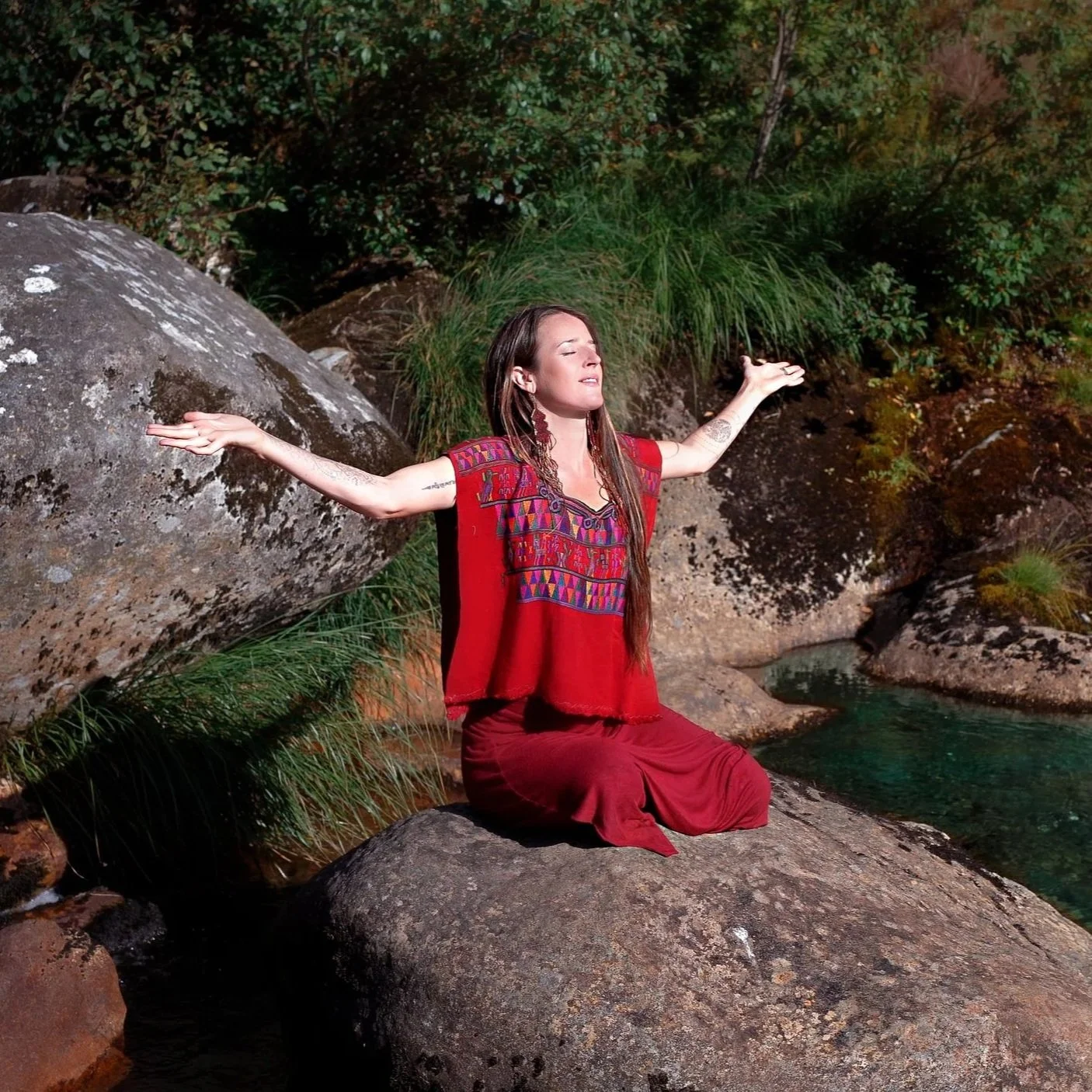 A woman in a red embroidered top and skirt kneels on a large rock by a stream with her arms outstretched and eyes closed, surrounded by greenery and rocks.