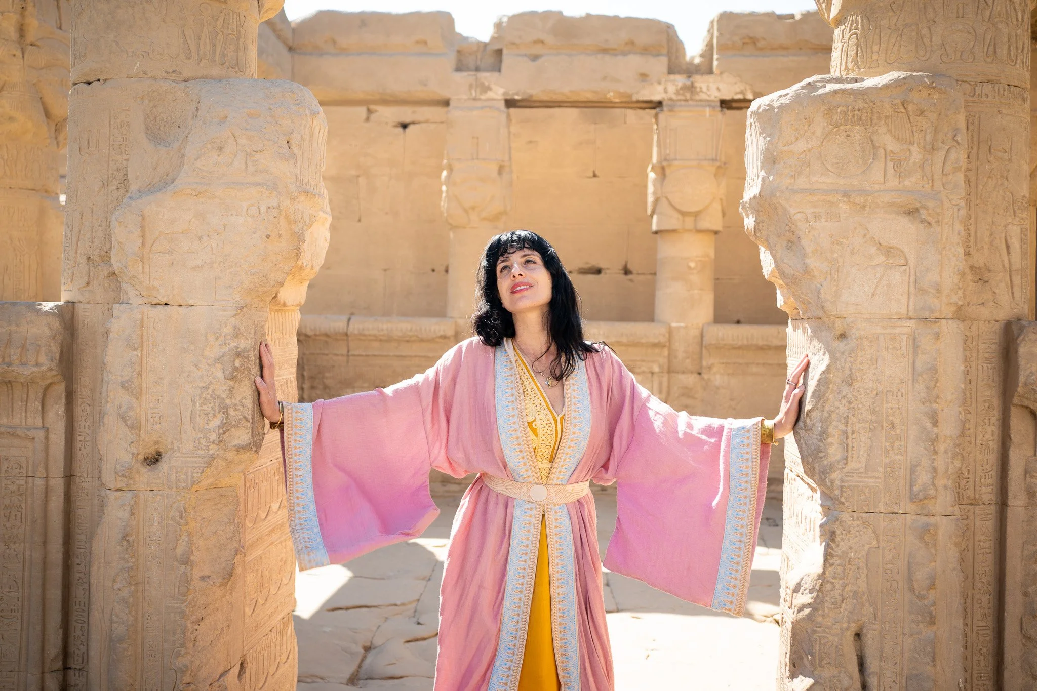 A woman in a pink and yellow flowing dress with wide sleeves stands between ancient Egyptian stone columns with hieroglyphics, gazing upward with her arms extended to touch the columns, in a desert-like ruins setting.