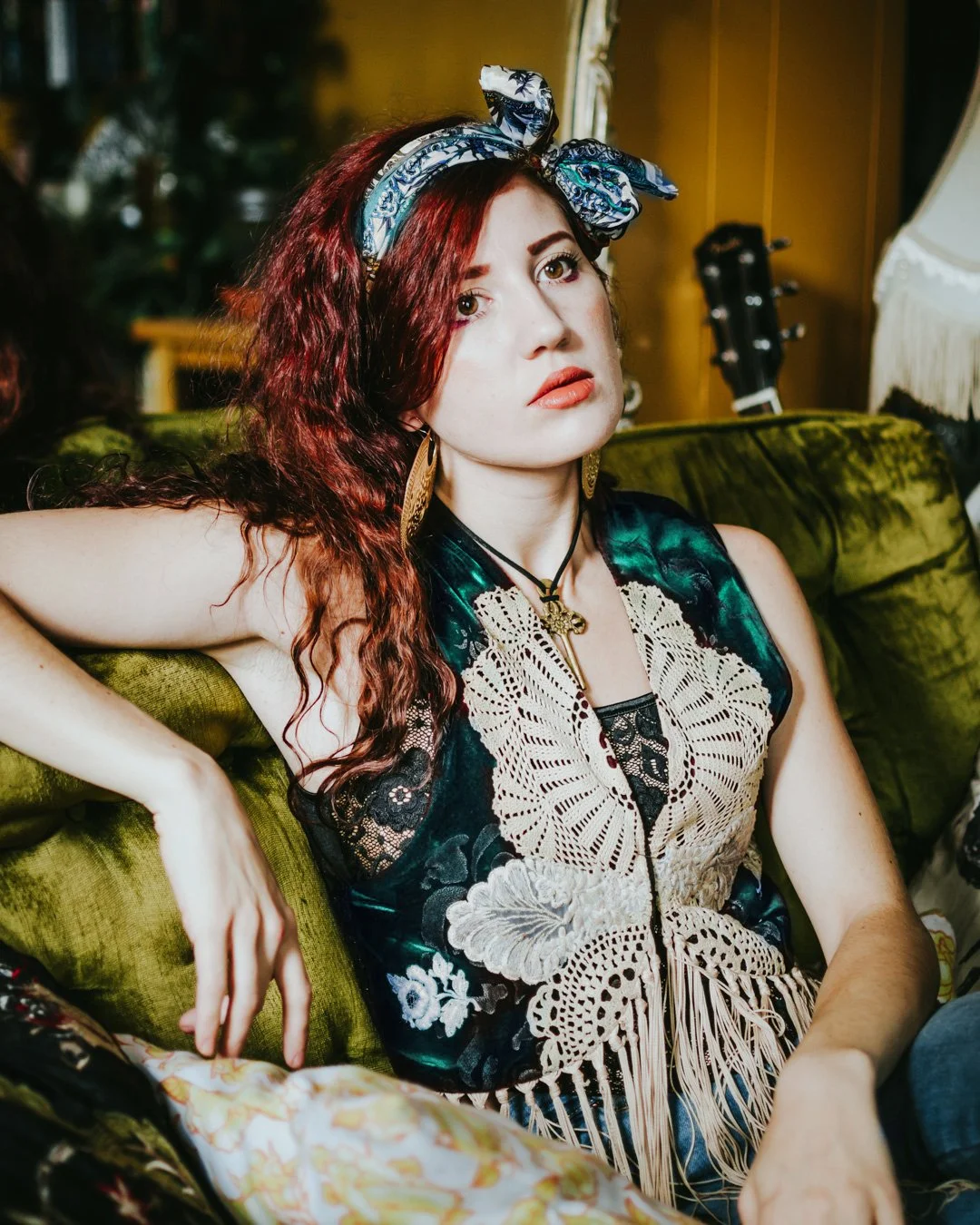 A young woman with red curly hair wearing a bandana, black and teal dress with lace details, gold jewelry, sitting on a green velvet couch.