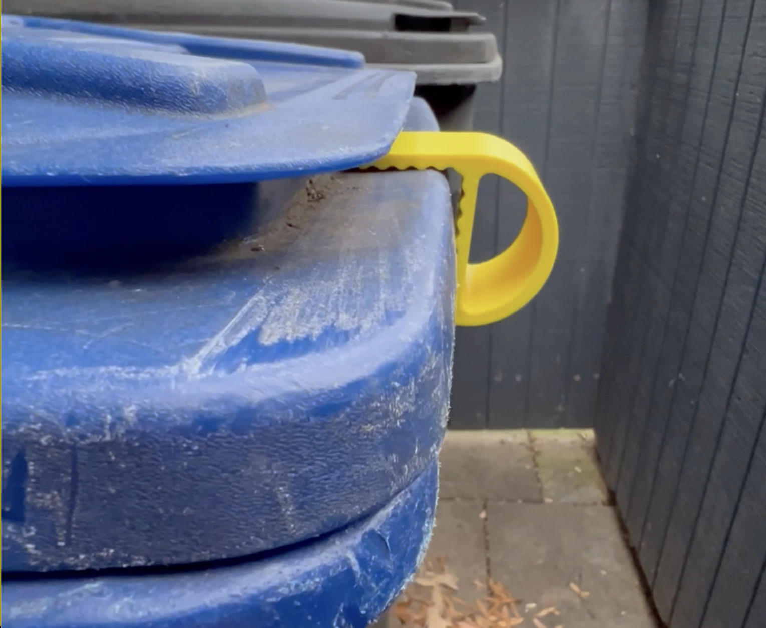 Close-up of blue plastic recycling bins with a yellow handle, stacked outdoors against a dark wooden fence.