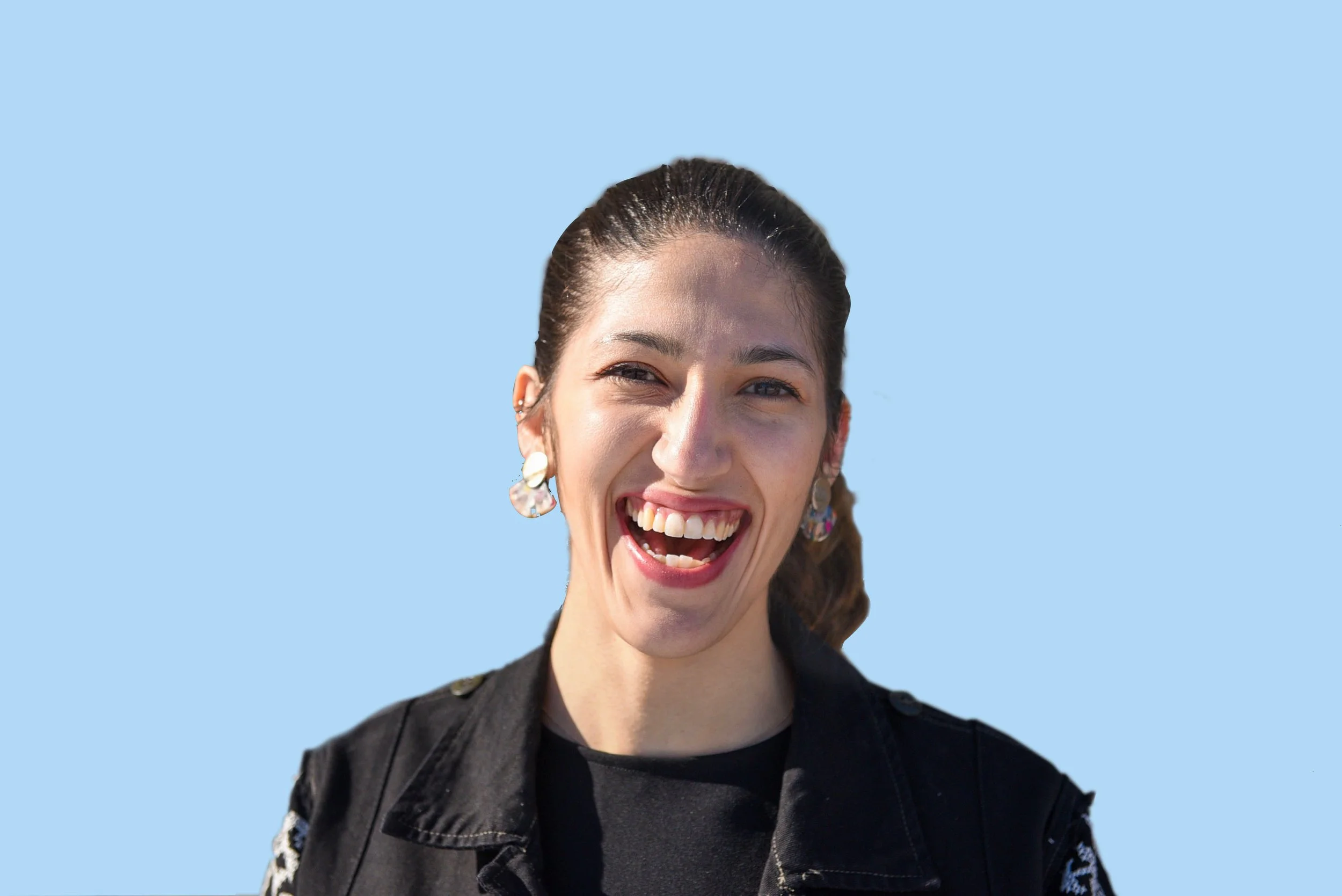 A young woman smiling broadly, showing her teeth, with dark hair tied back, wearing a black jacket and colorful earrings, against a light blue background.