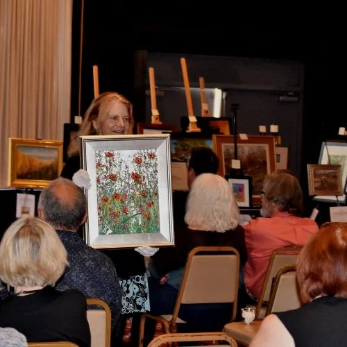 A woman in white gloves presenting a framed painting of red and orange flowers in a field to an audience at an art auction or gallery event.