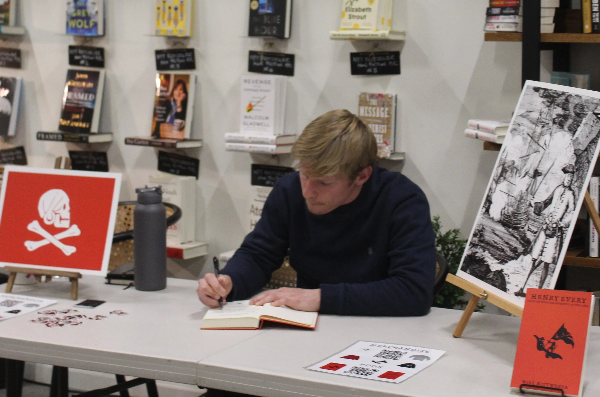 A man with blond hair wearing a dark sweater, sitting at a table signing a book during a book signing event. The table has promotional materials, including a large black and white illustration on an easel, a red sign with a skull and crossbones, and some smaller items. Bookshelves with various books are visible in the background.