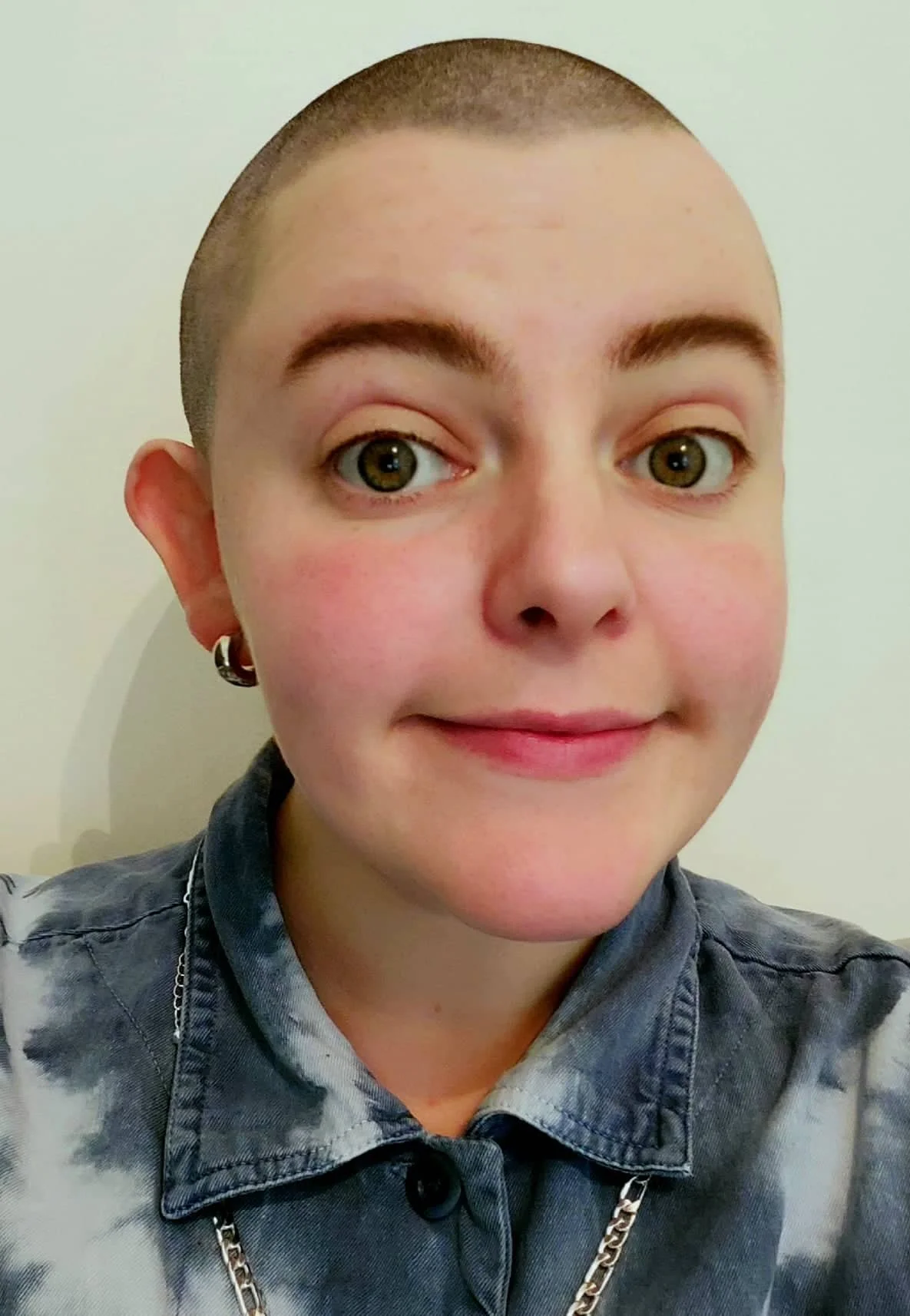 a headshot of an androgynous person with a shaved head. They have large dark green eyes and are smiling at the camera. They have a thick silver hoop earing in their right ear.