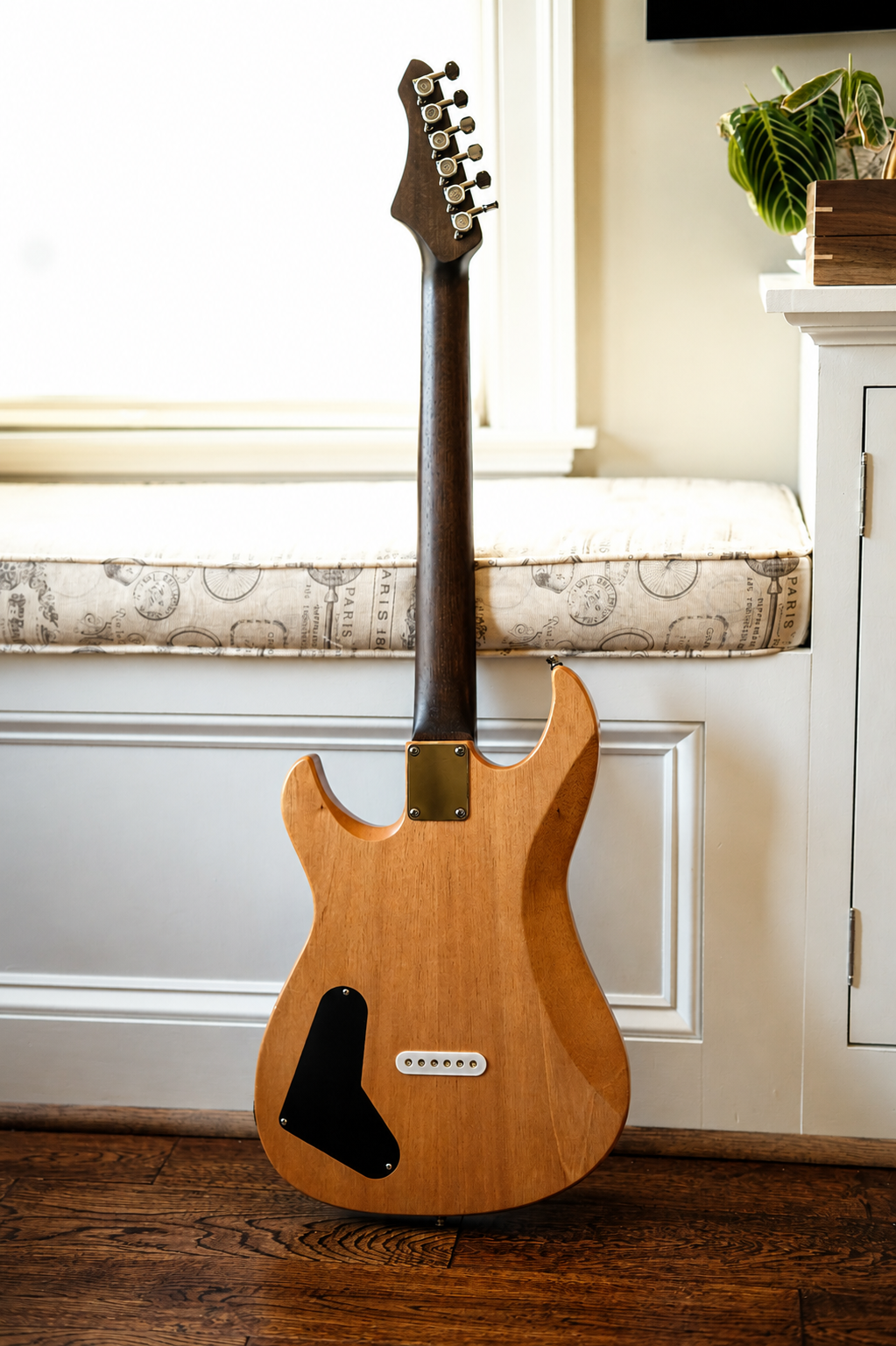An unfinished electric guitar with a natural wood finish, leaning against a white bench with a cushion, next to a white cabinet and a potted plant.