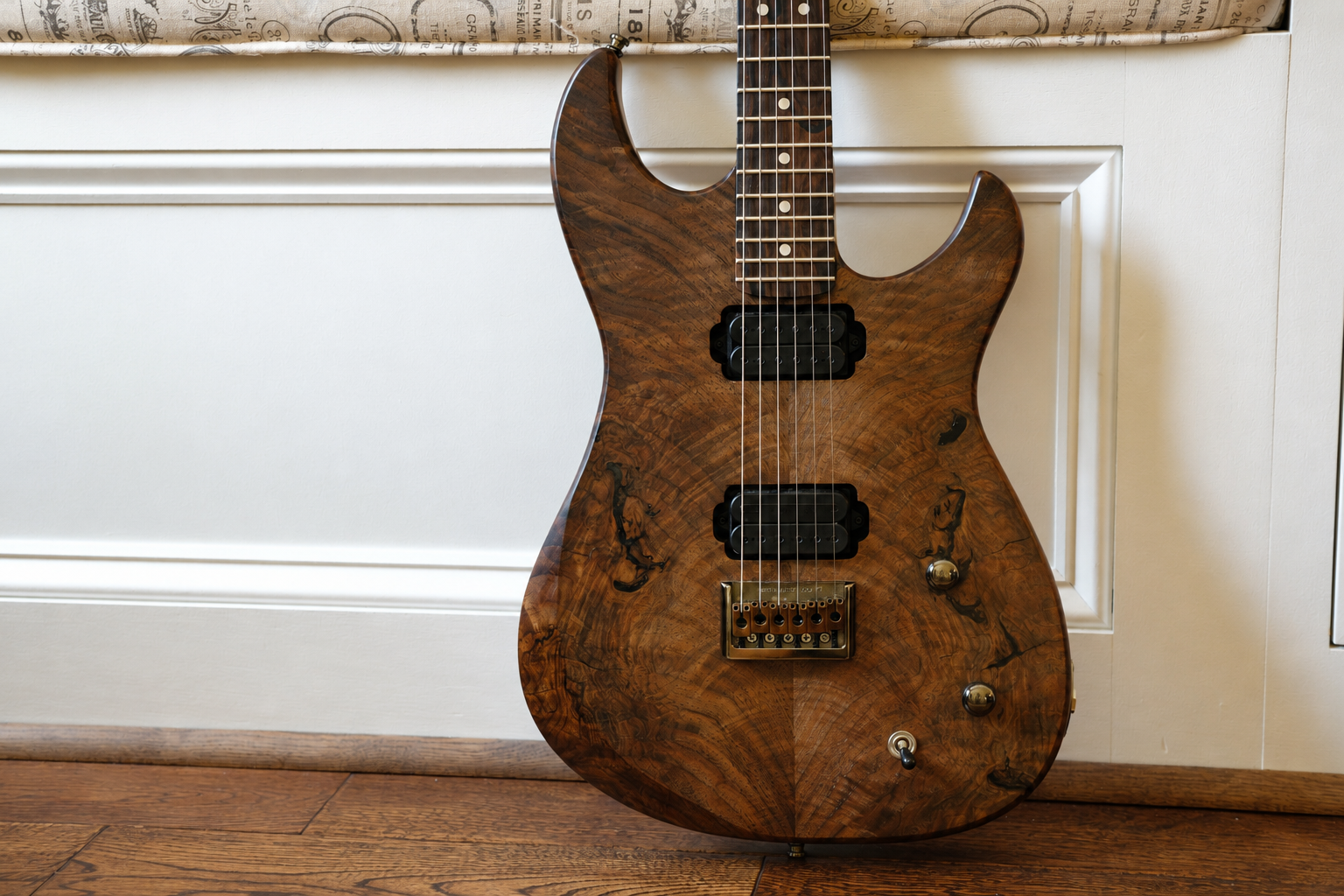 A wooden electric guitar with a natural finish, two pickups, a bridge, and control knobs, leaning against a white wall with a wood floor.