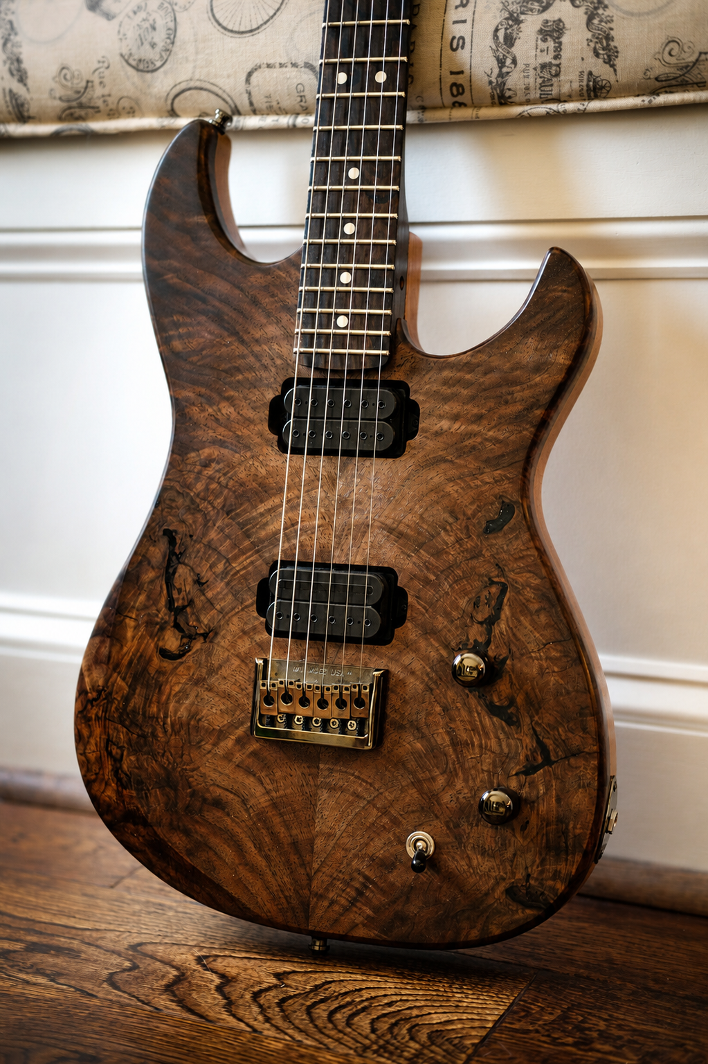 A wooden electric guitar with a distinctive natural grain finish, black pickups, and gold hardware, leaning against a white wall near a window with decorative paper trim.