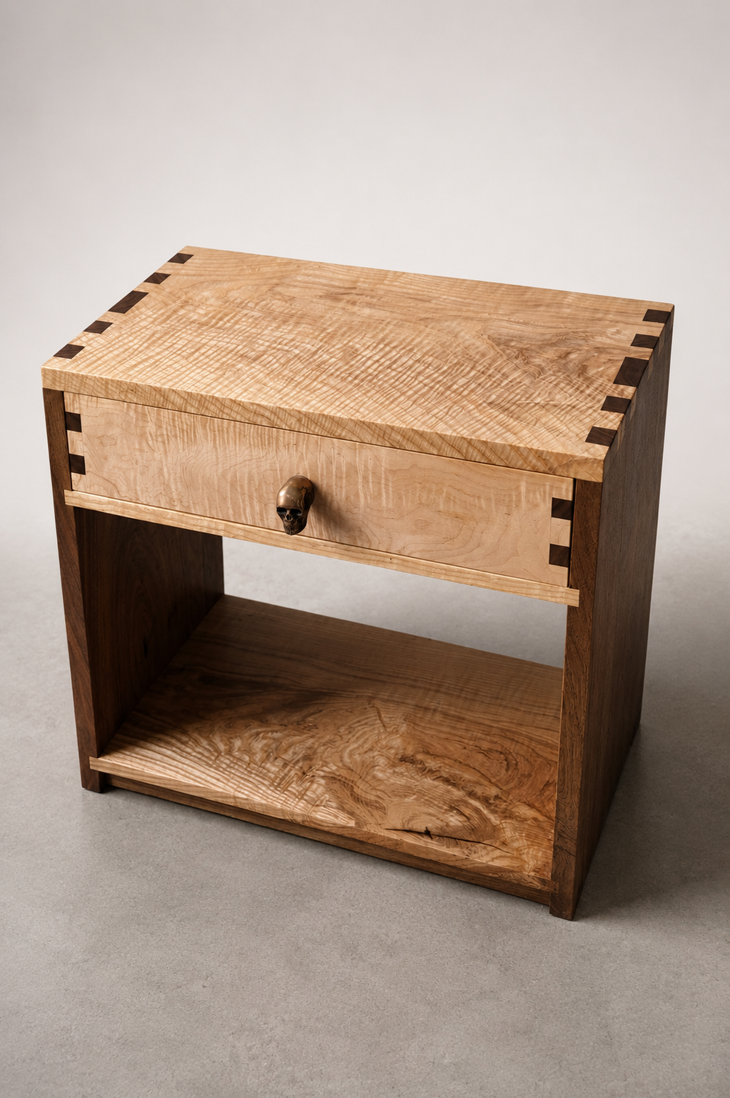 A wooden nightstand with a single drawer and a gold skull-shaped handle, featuring dovetail joints, placed on a gray carpeted floor against a plain white background.