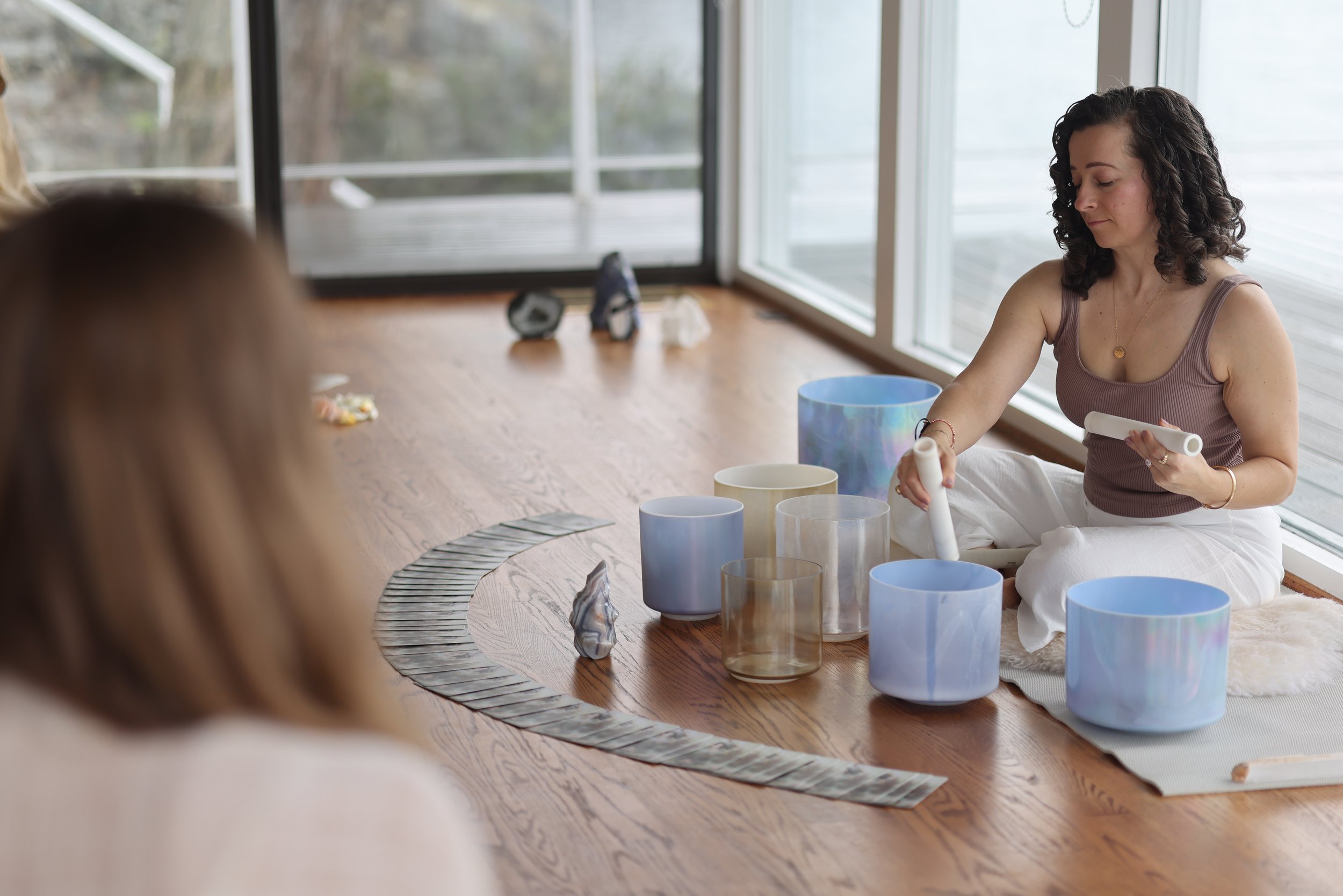A woman sitting cross-legged on a rug, playing crystal singing bowls and chimes, with a second person blurred in the foreground, inside a room with large windows and a view outside.