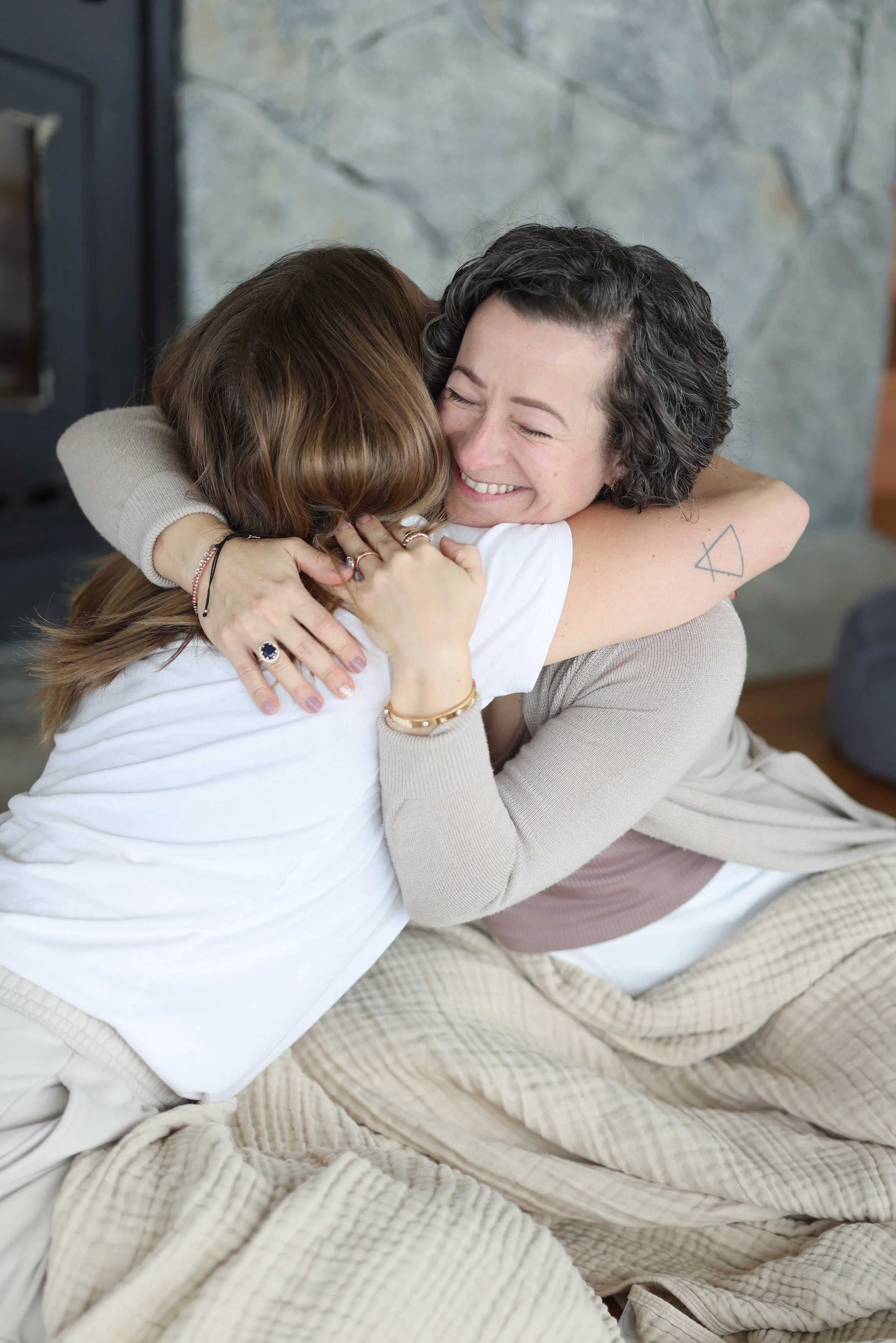 Two women hugging and smiling warmly in a cozy indoor setting with a stone fireplace in the background.