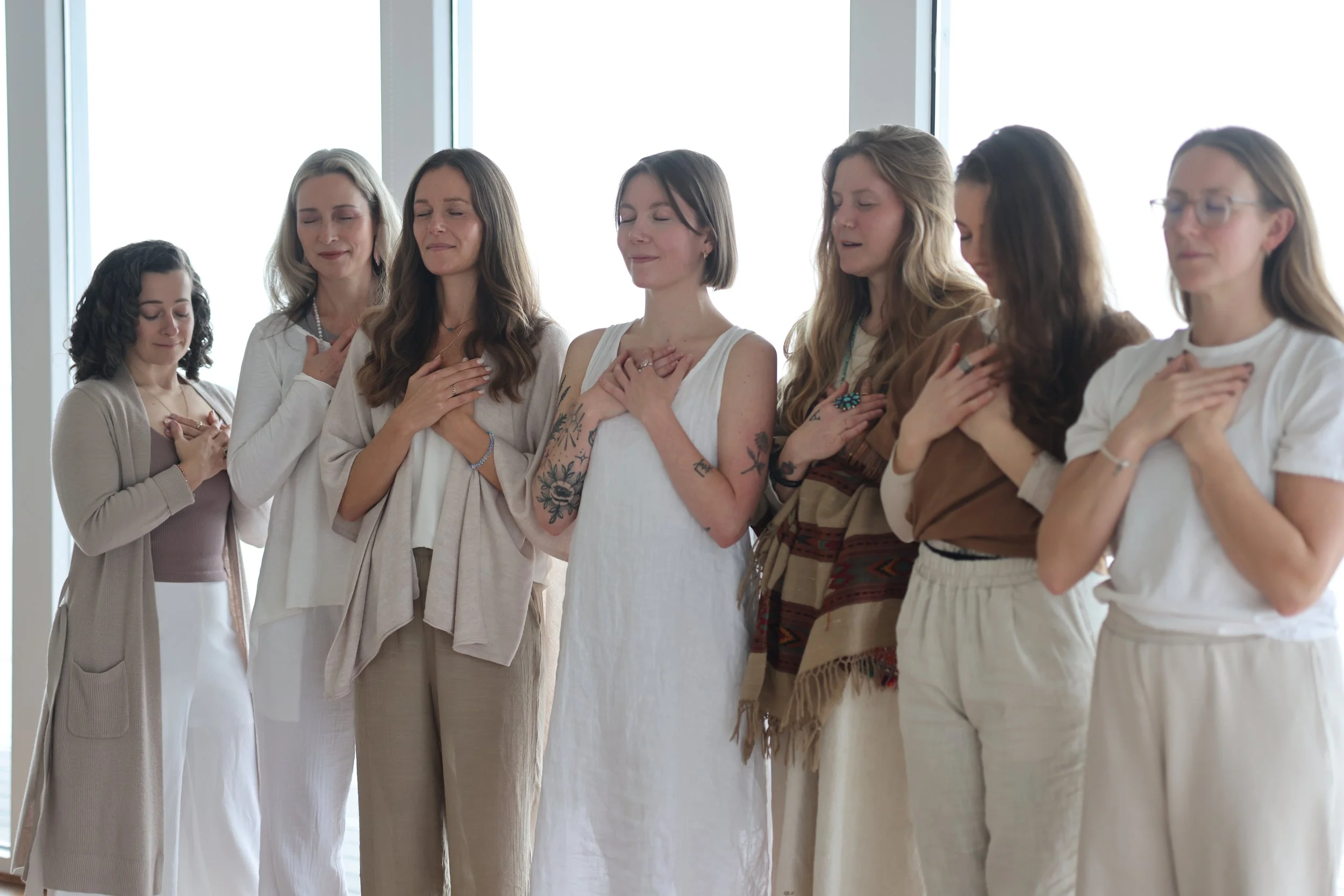 Seven women standing together with their hands on their chests, eyes closed, appearing to be praying or meditating in a bright indoor setting.