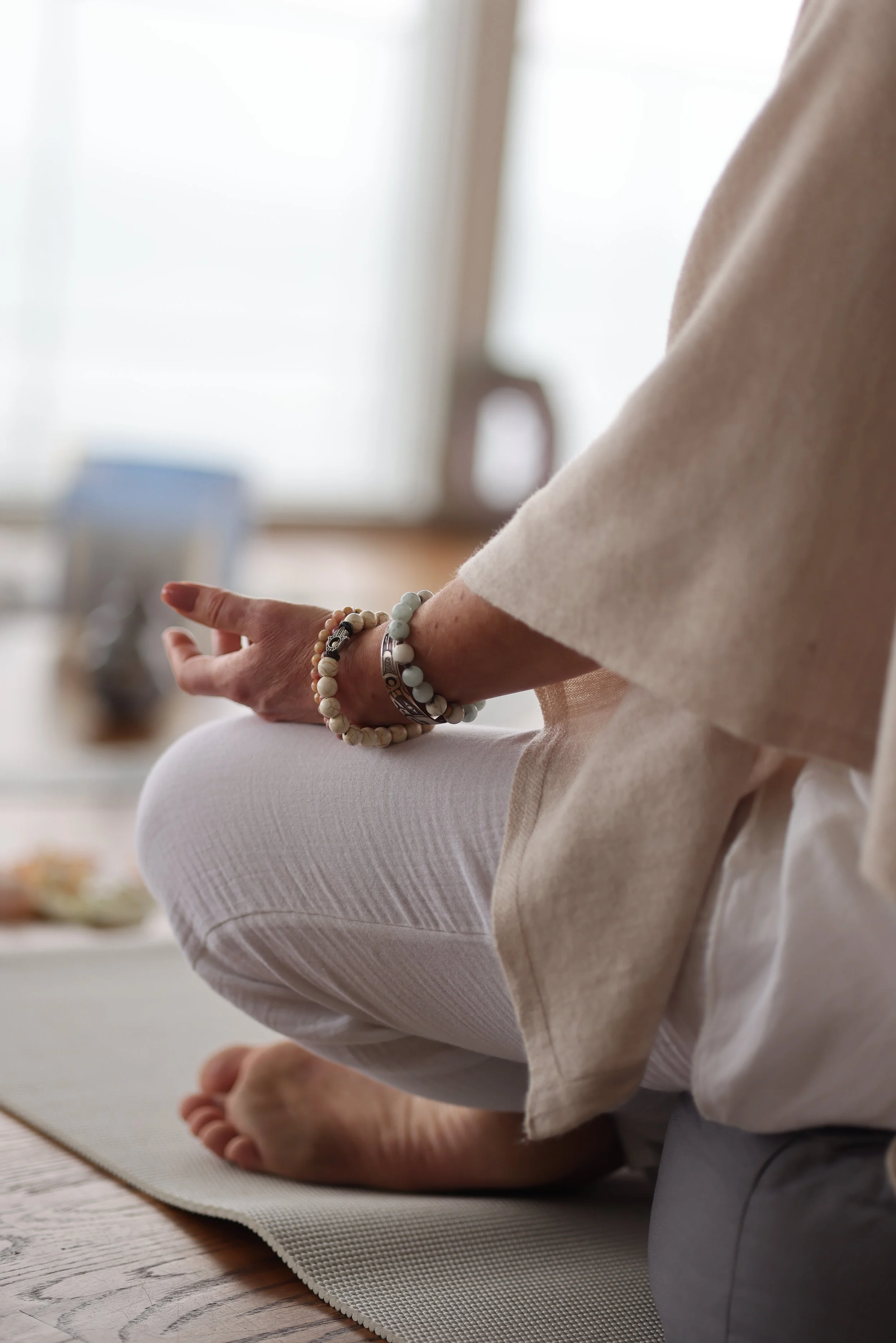 Person practicing meditation or yoga on a mat in a bright room, sitting cross-legged with hand resting on knee, wearing bracelets and light-colored clothing.