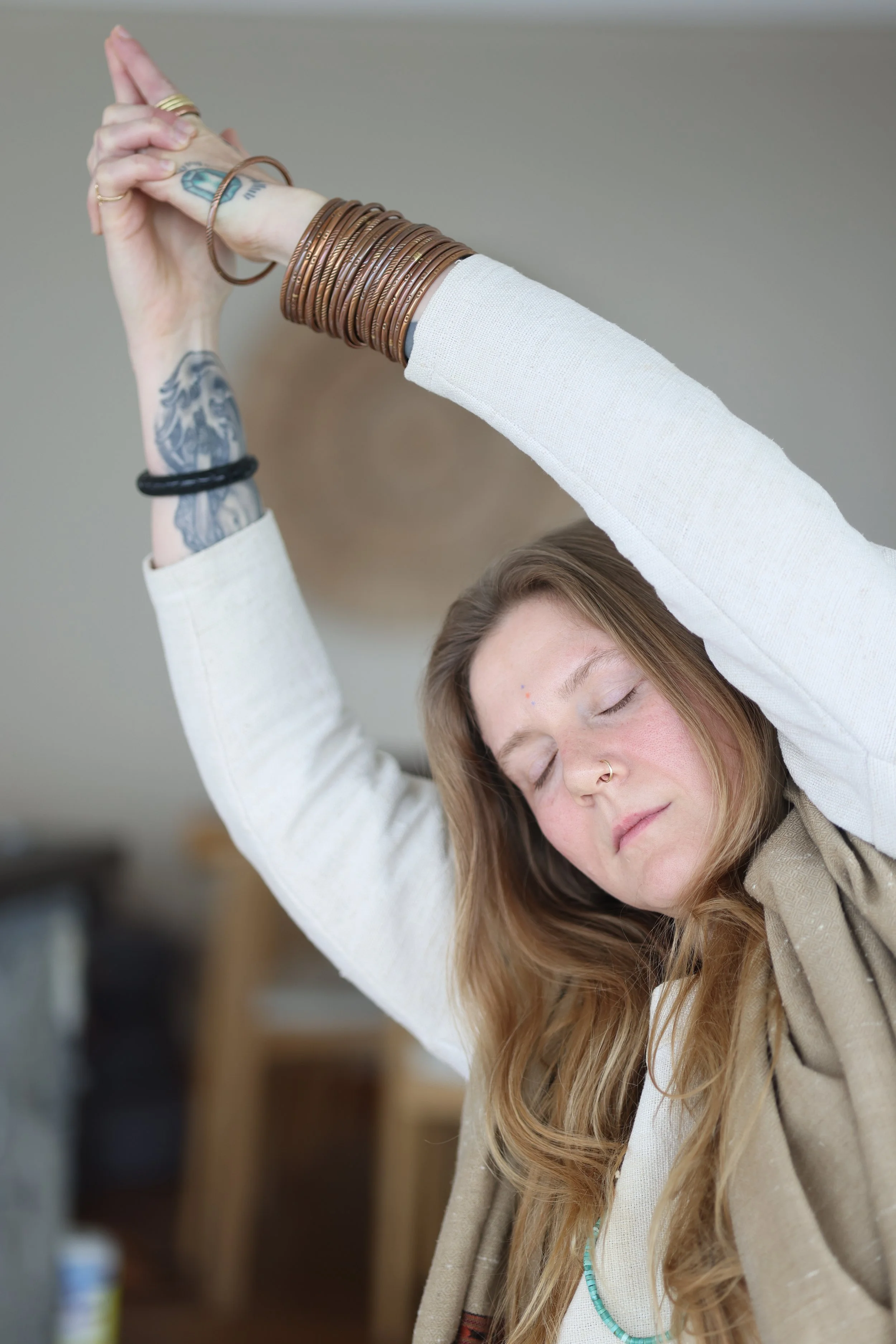 A woman with long hair, tattoos, and multiple bracelets is stretching with her arms raised above her head and her eyes closed.