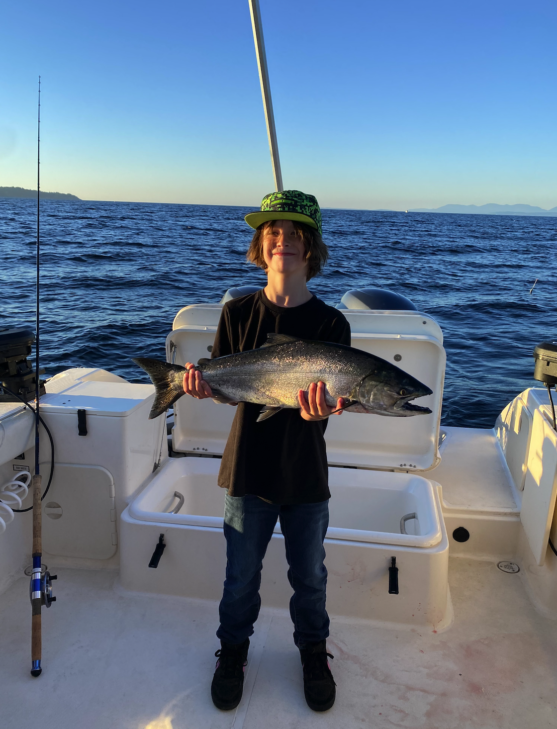 A young boy on a boat holding a large fish, with the ocean and distant land in the background during sunset.