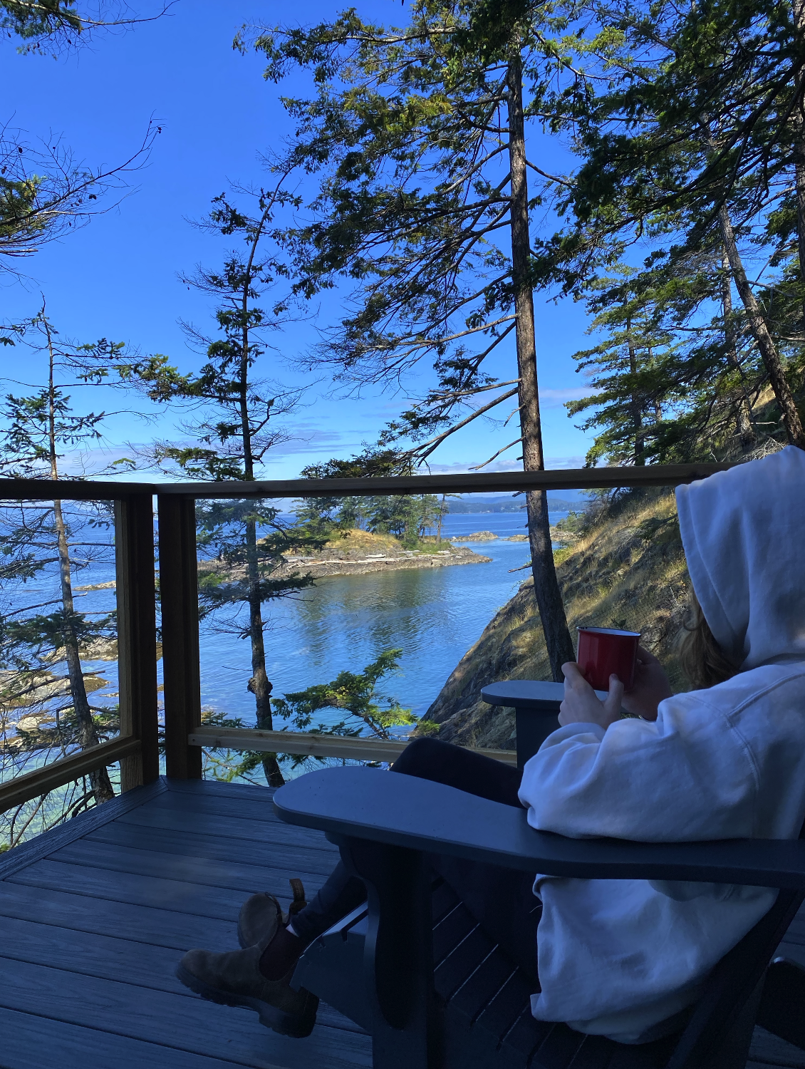 Person in a white hoodie sitting on a porch overlooking a coastal landscape with trees, water, and distant islands, holding a red mug.