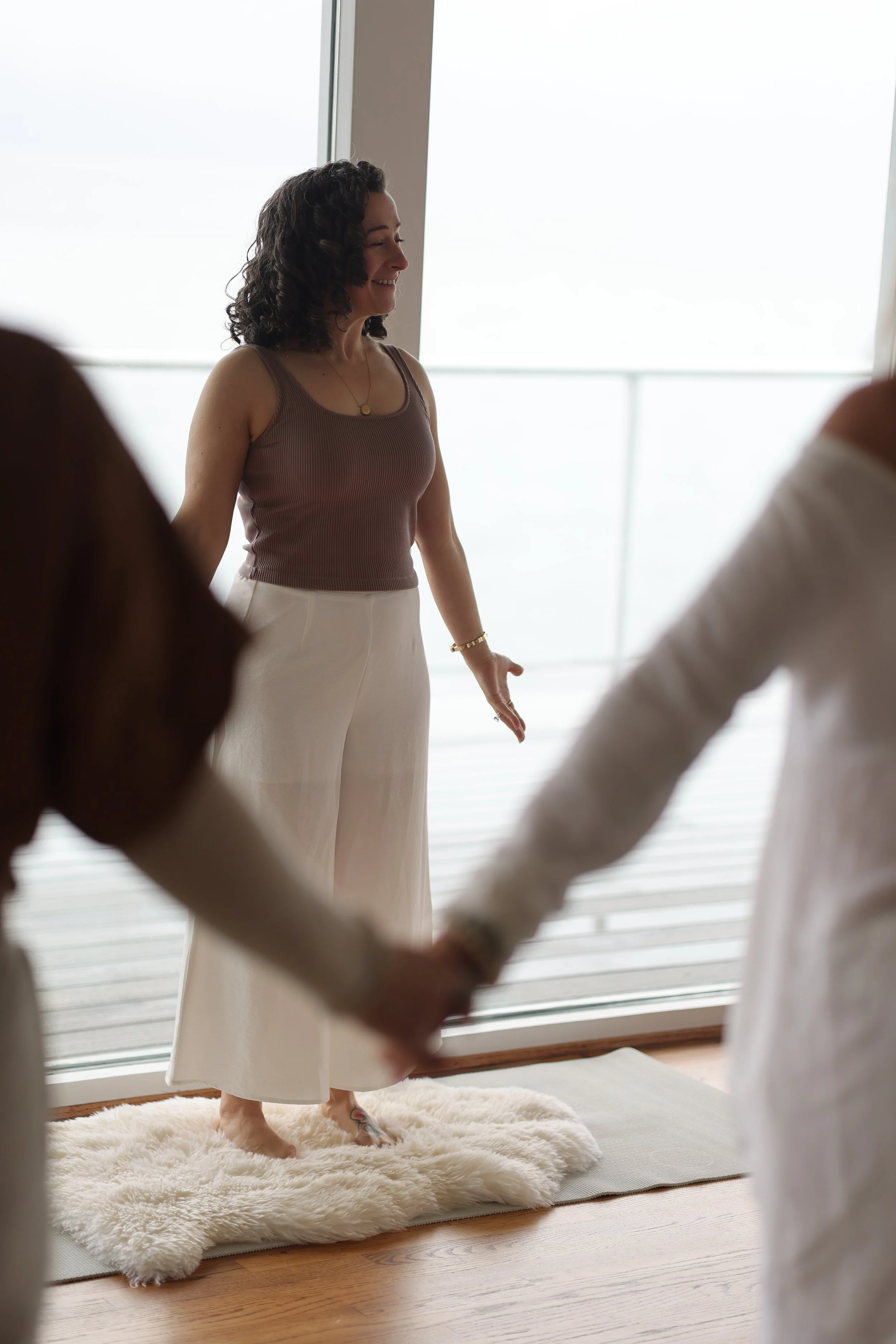 Woman standing on a white fluffy rug holding hands with two other people during a gathering or ceremony in front of large windows.
