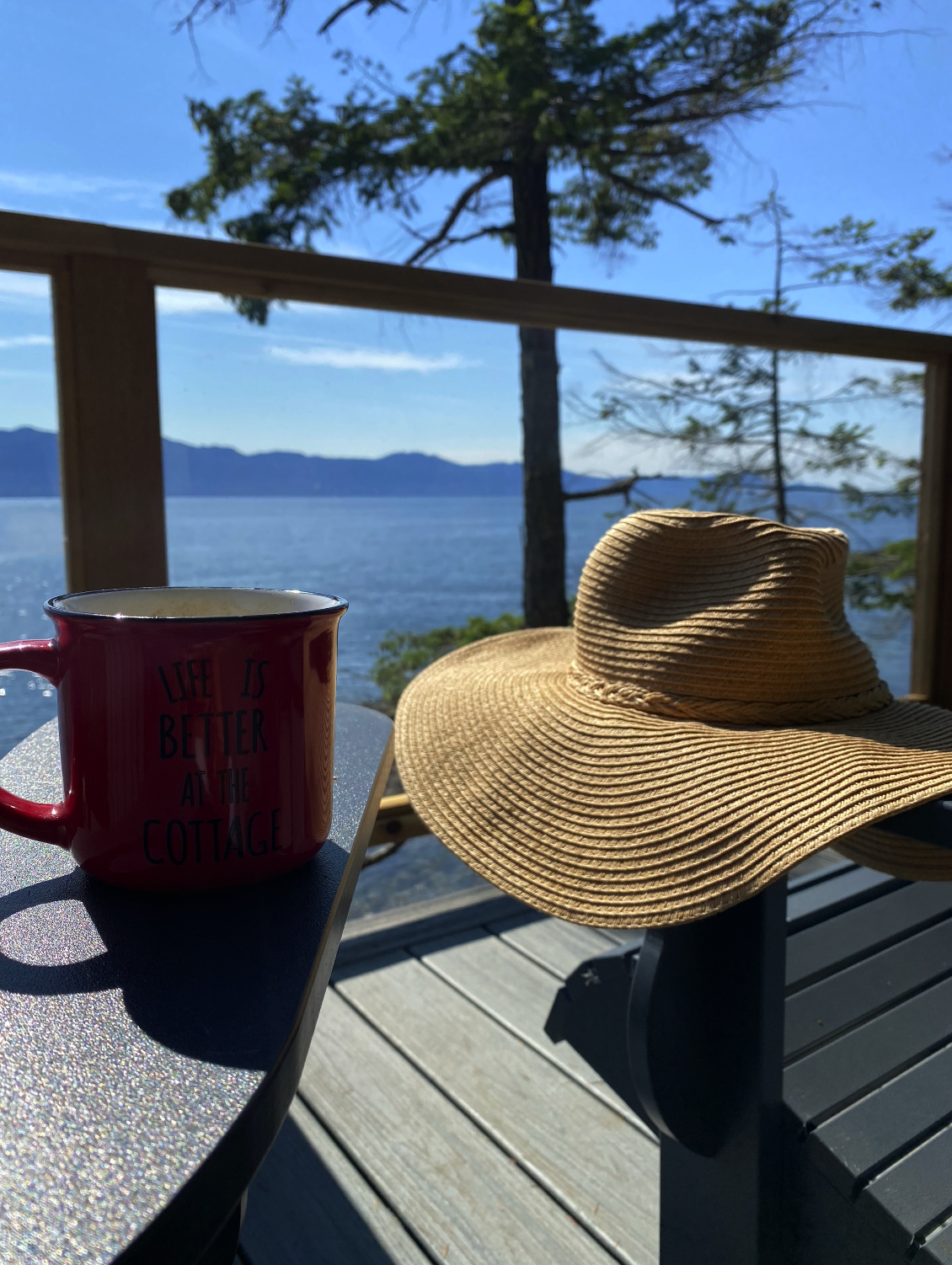 A red mug with the words 'Life is better at the cottage' and a wide-brimmed straw hat resting on a black bench on a wooden deck overlooking a lake, with trees and mountains in the background on a sunny day.