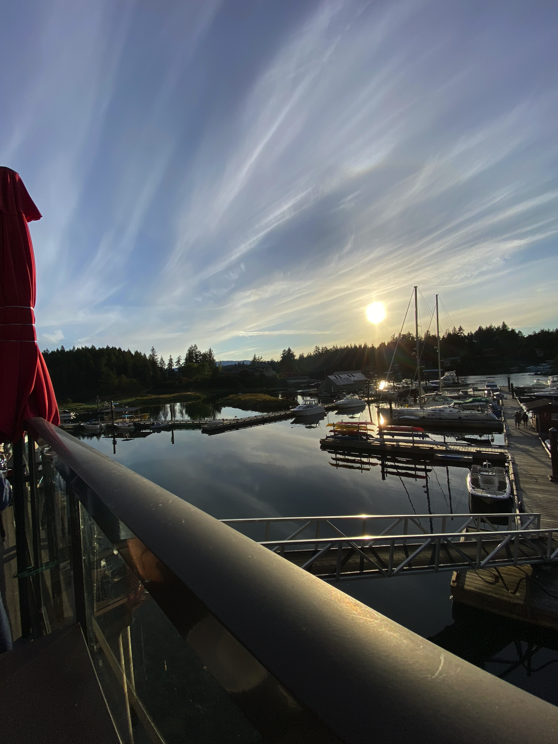 Sunset over a marina with boats docked, calm water reflecting the sky, trees in the background, and part of a boat's railing in the foreground.