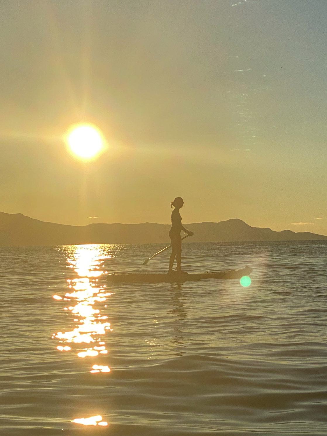 A person paddleboarding on calm water during sunset with mountains in the background.